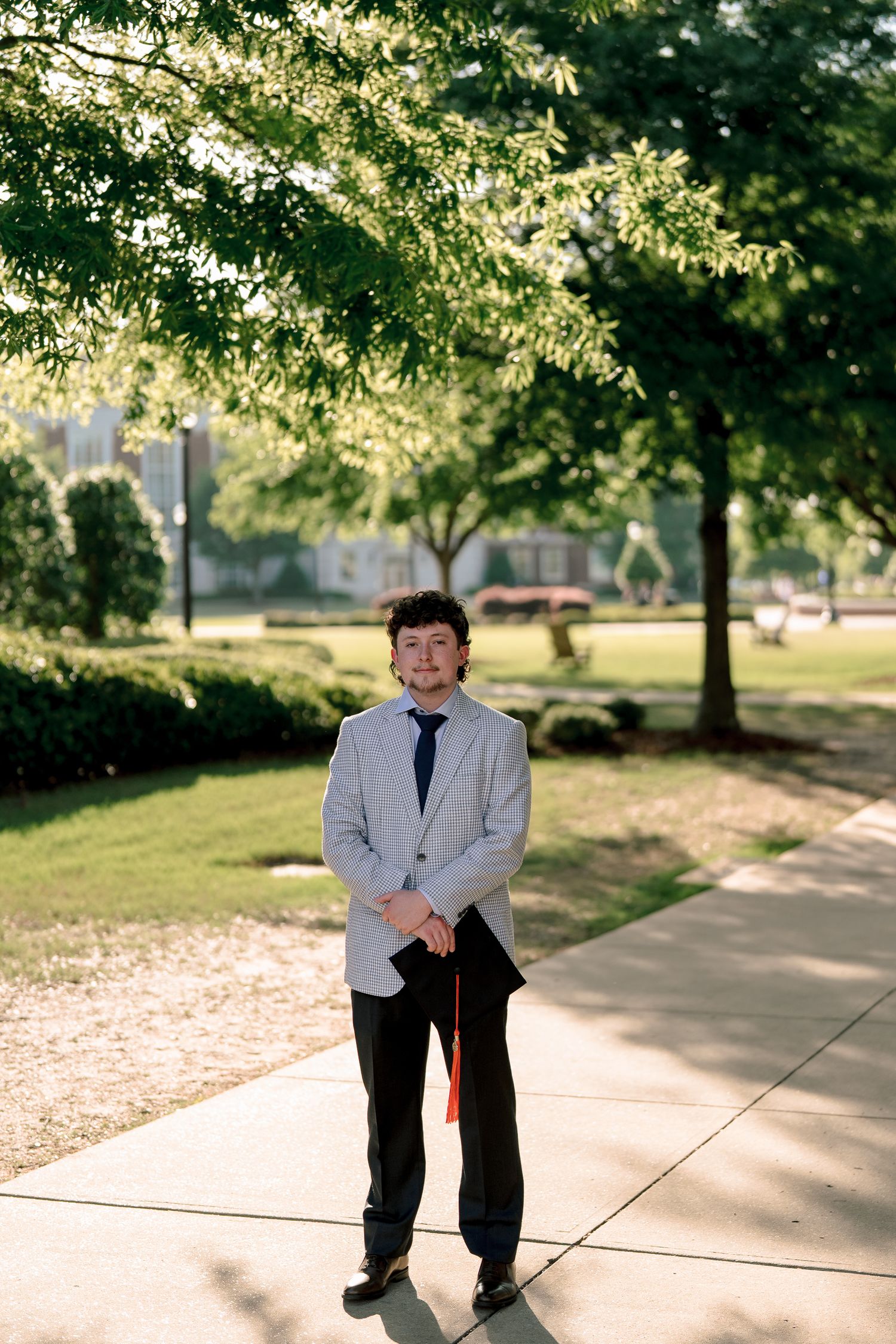 Young man in striped blazer and tie standing on campus sidewalk surrounded by trees.