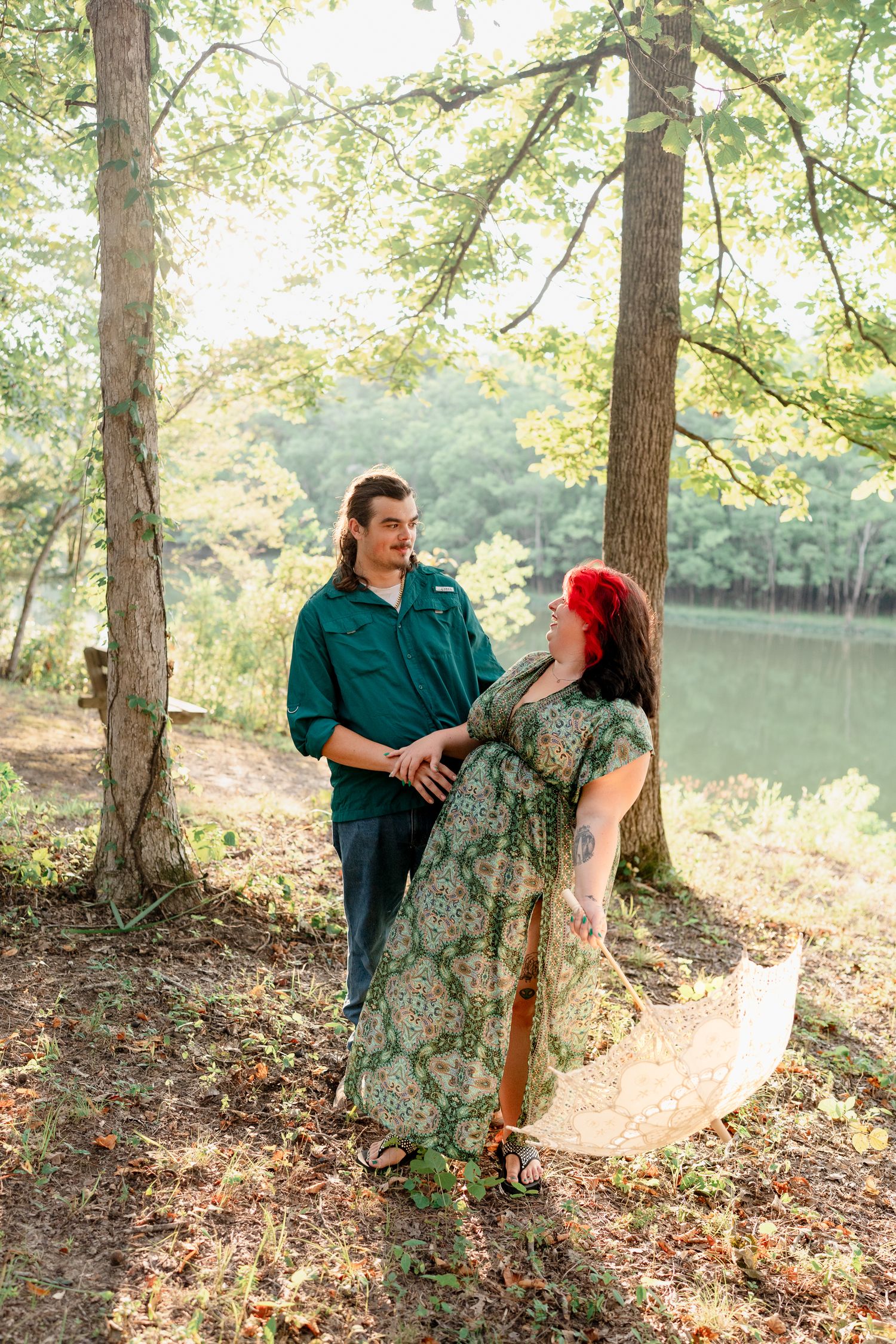 A couple poses playfully near a lake with sunlit trees creating a romantic woodland setting.