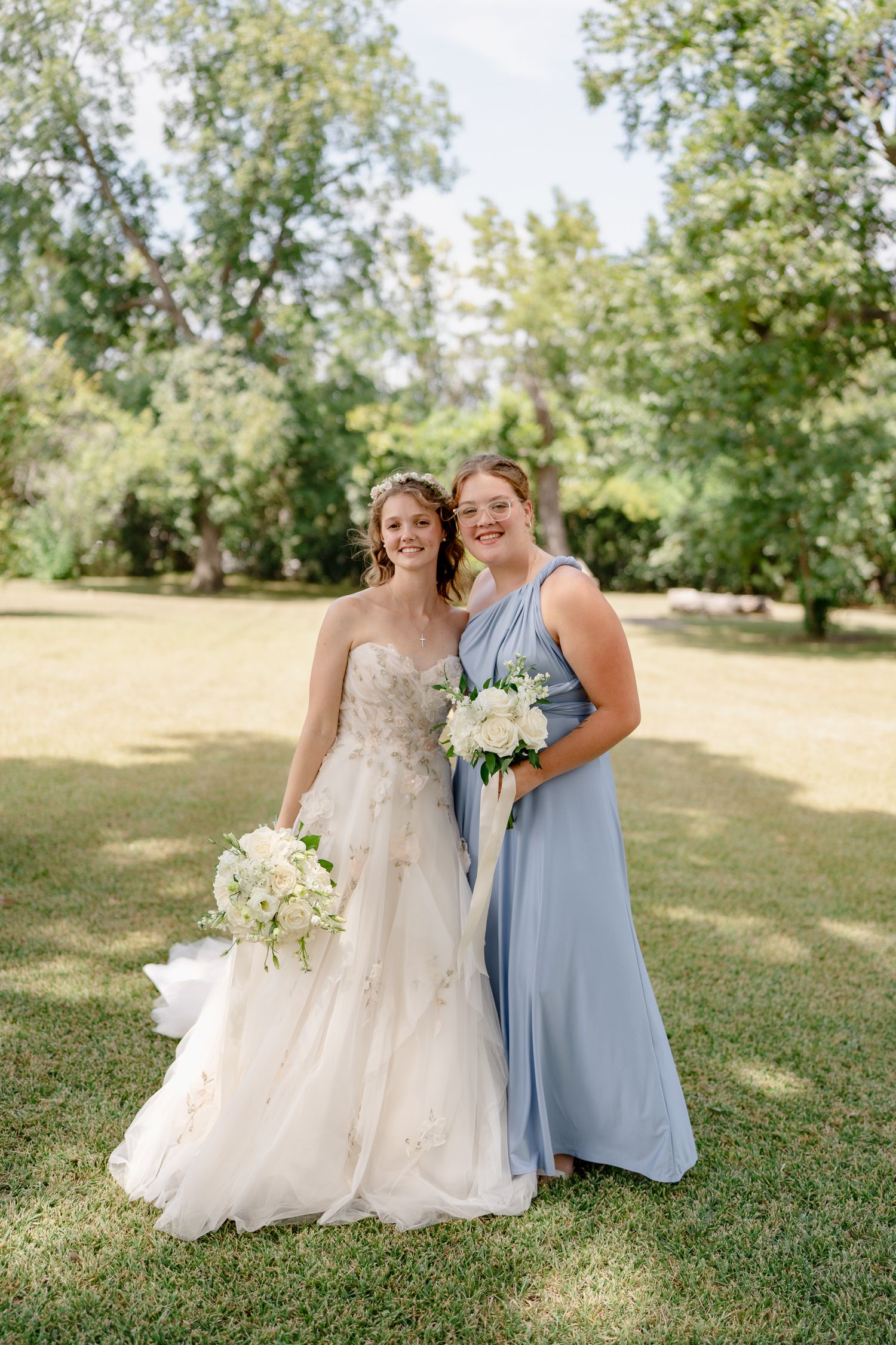 Two people pose together outdoors, one wearing a white wedding gown and one in a powder blue dress with floral bouquets.