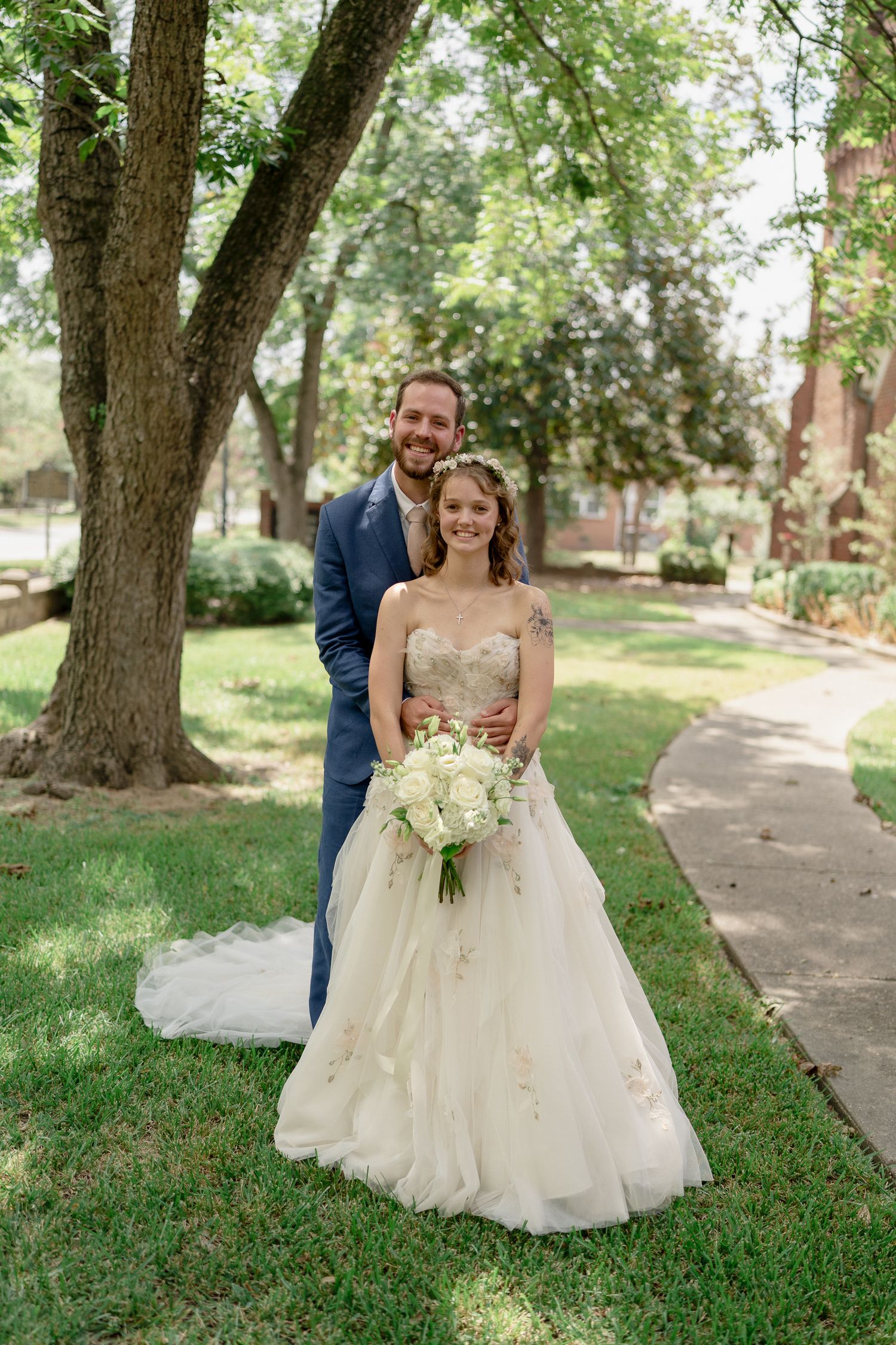 A couple walks along a tree-lined sidewalk on their wedding day, with the bride wearing a flowing white gown.