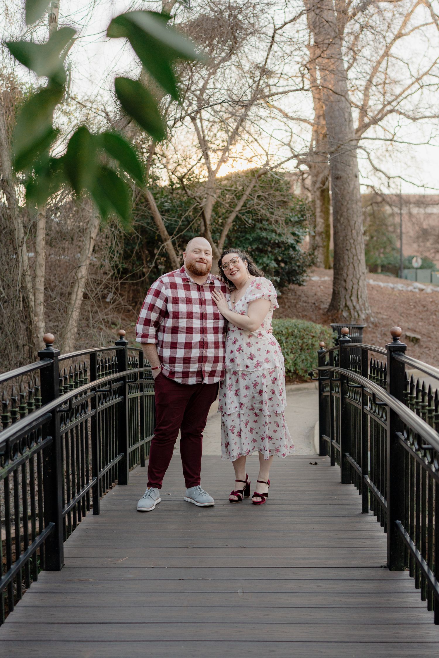 A couple stands together on a wooden bridge in a park, wearing coordinating plaid and floral outfits.