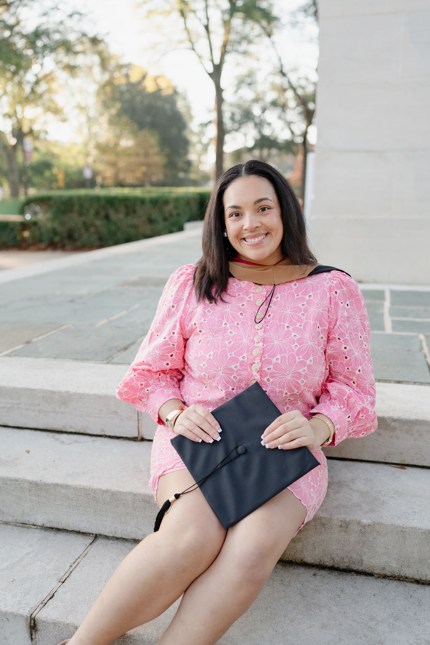A graduate in pink dress sits on concrete steps holding a graduation cap.