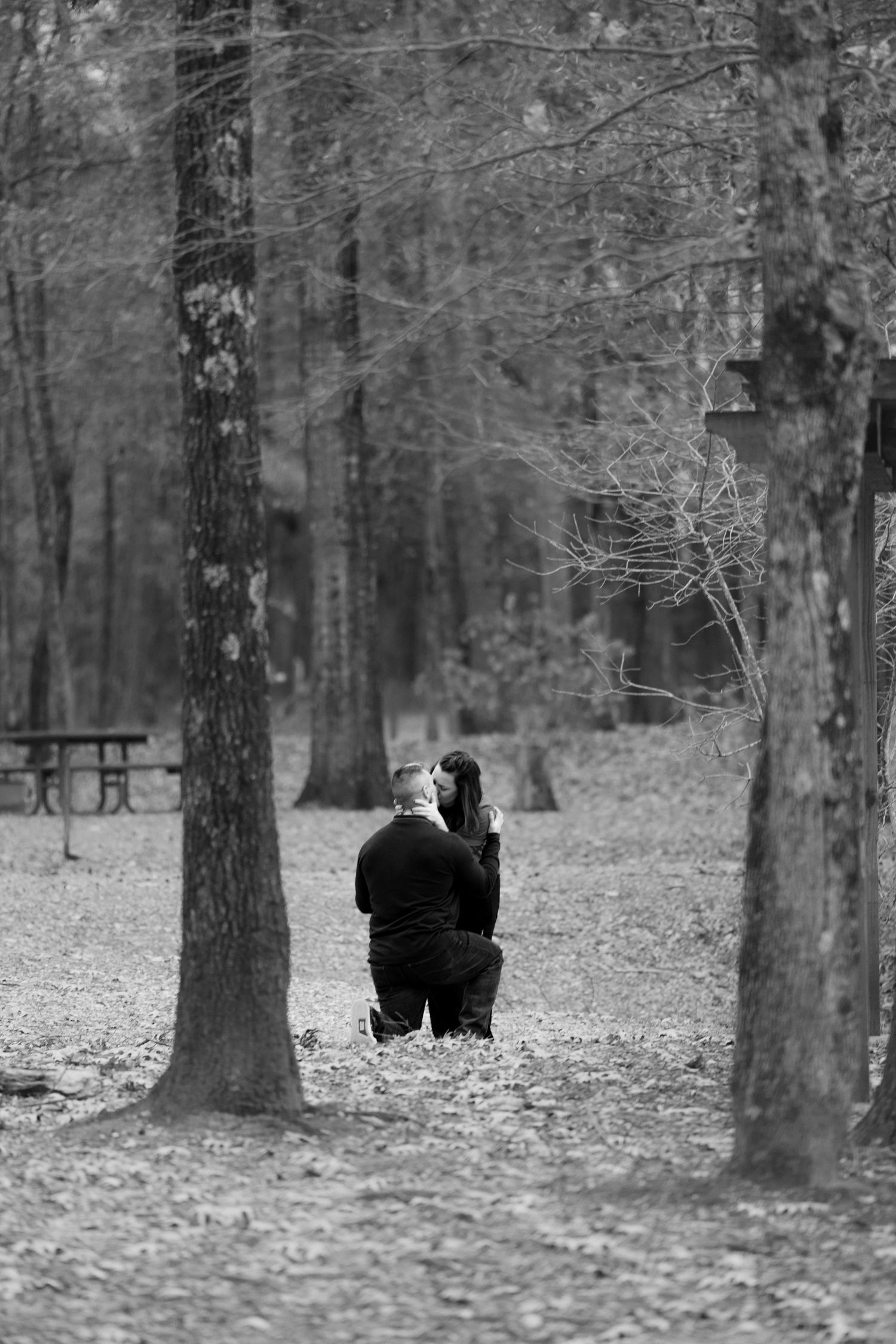 A couple walks through a wooded park with bare trees and fallen leaves on a winter day.