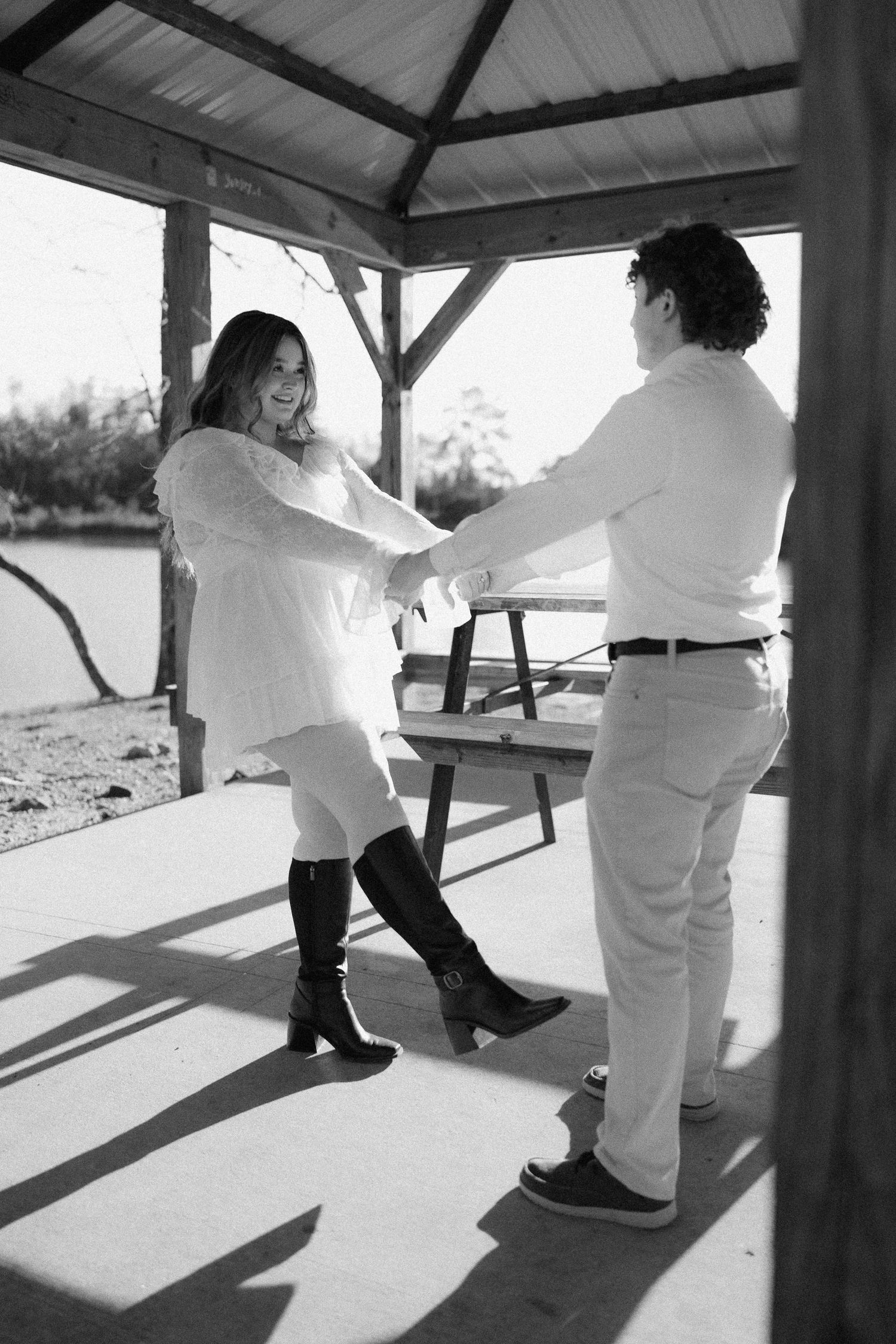 Black and white photo of couple dancing playfully under a rustic wooden gazebo.