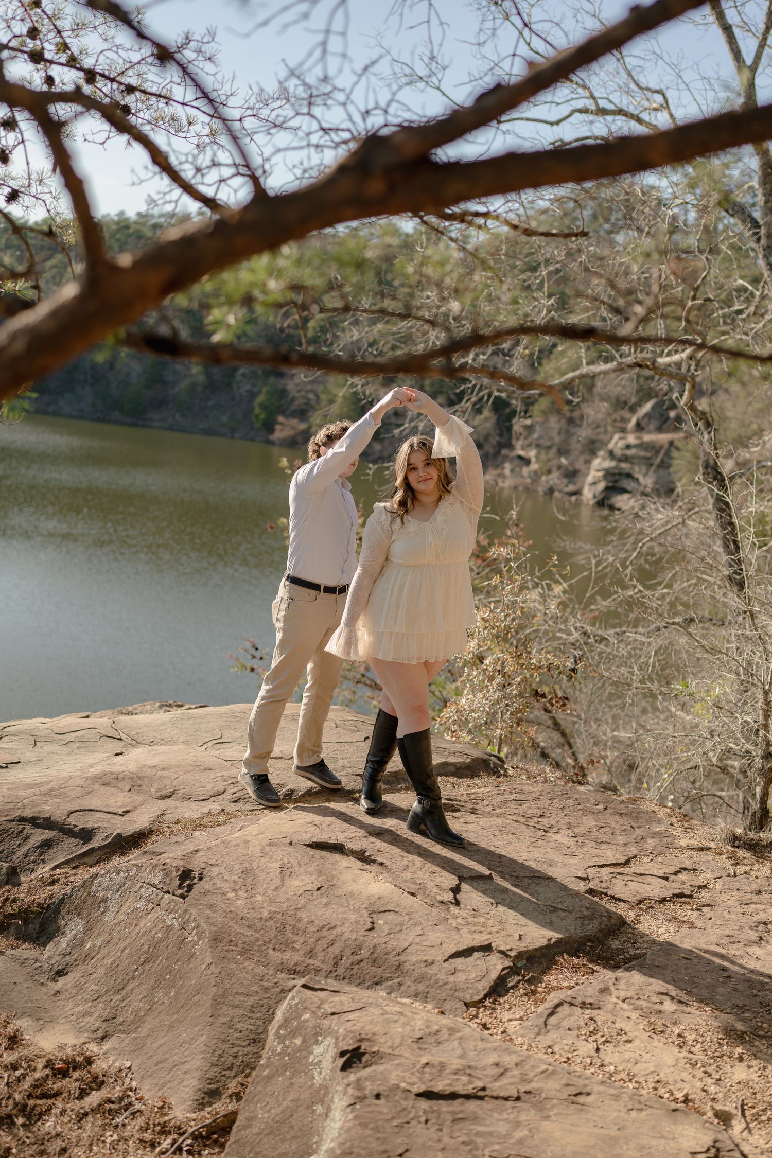 Couple dancing on rocky overlook by a lake, woman in white dress and black boots, man in khaki pants.
