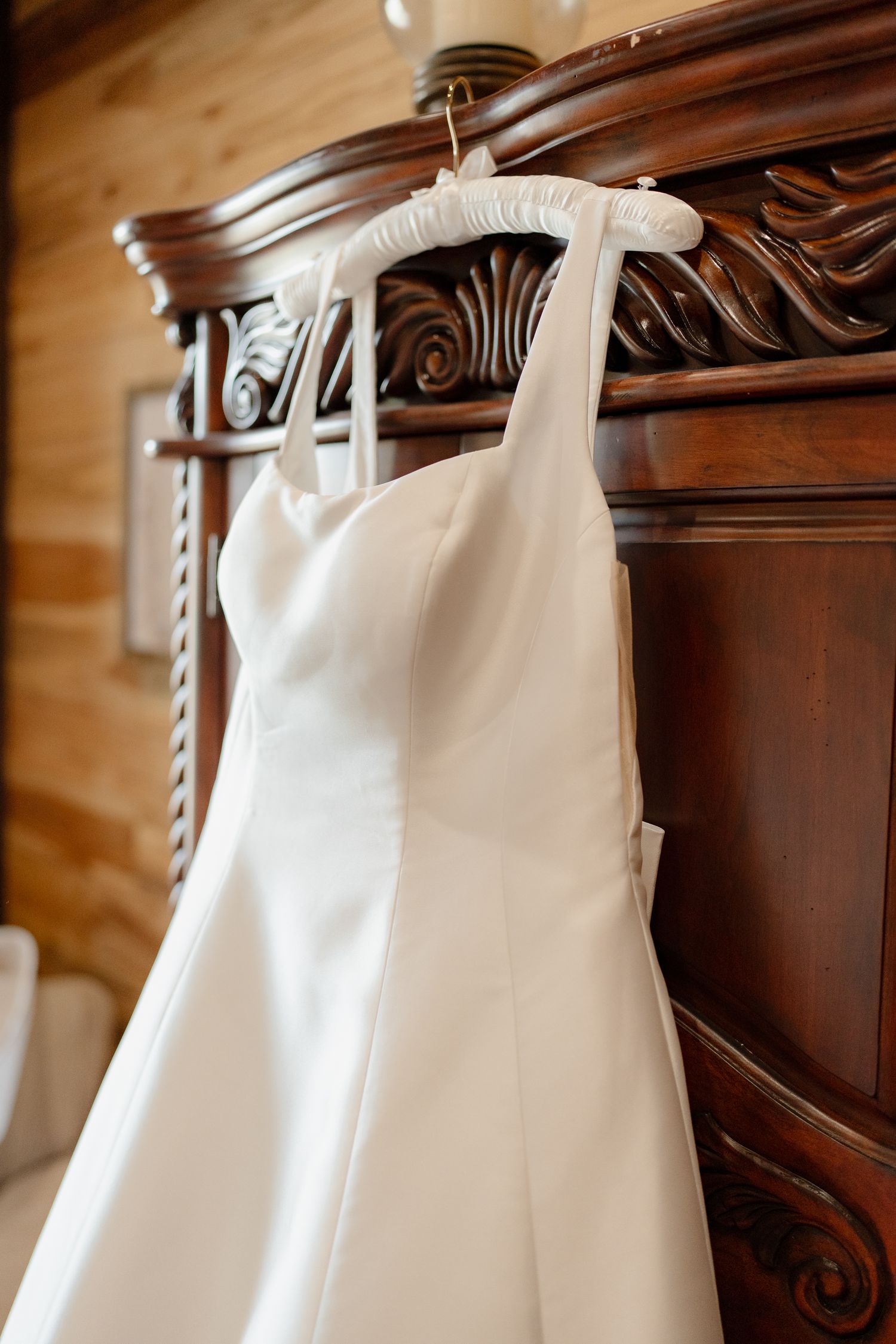 White wedding dress hanging on ornate wooden headboard in warm natural lighting.