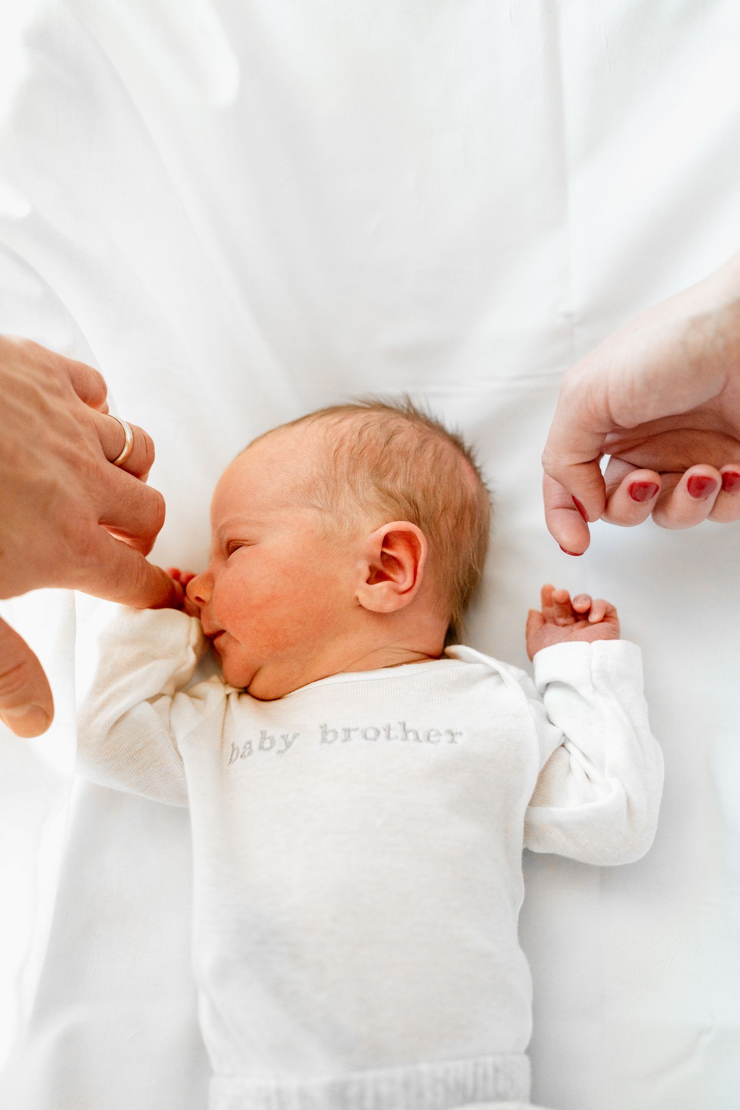 Sleeping baby in white 'baby brother' outfit on crisp white bedding.