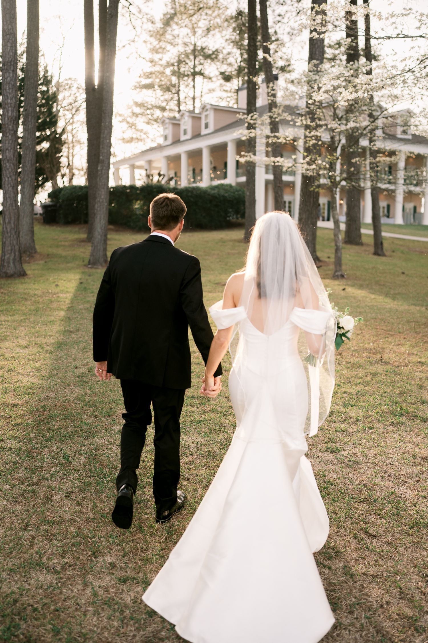 Newlyweds walking hand in hand towards stately mansion through tree-lined grounds.