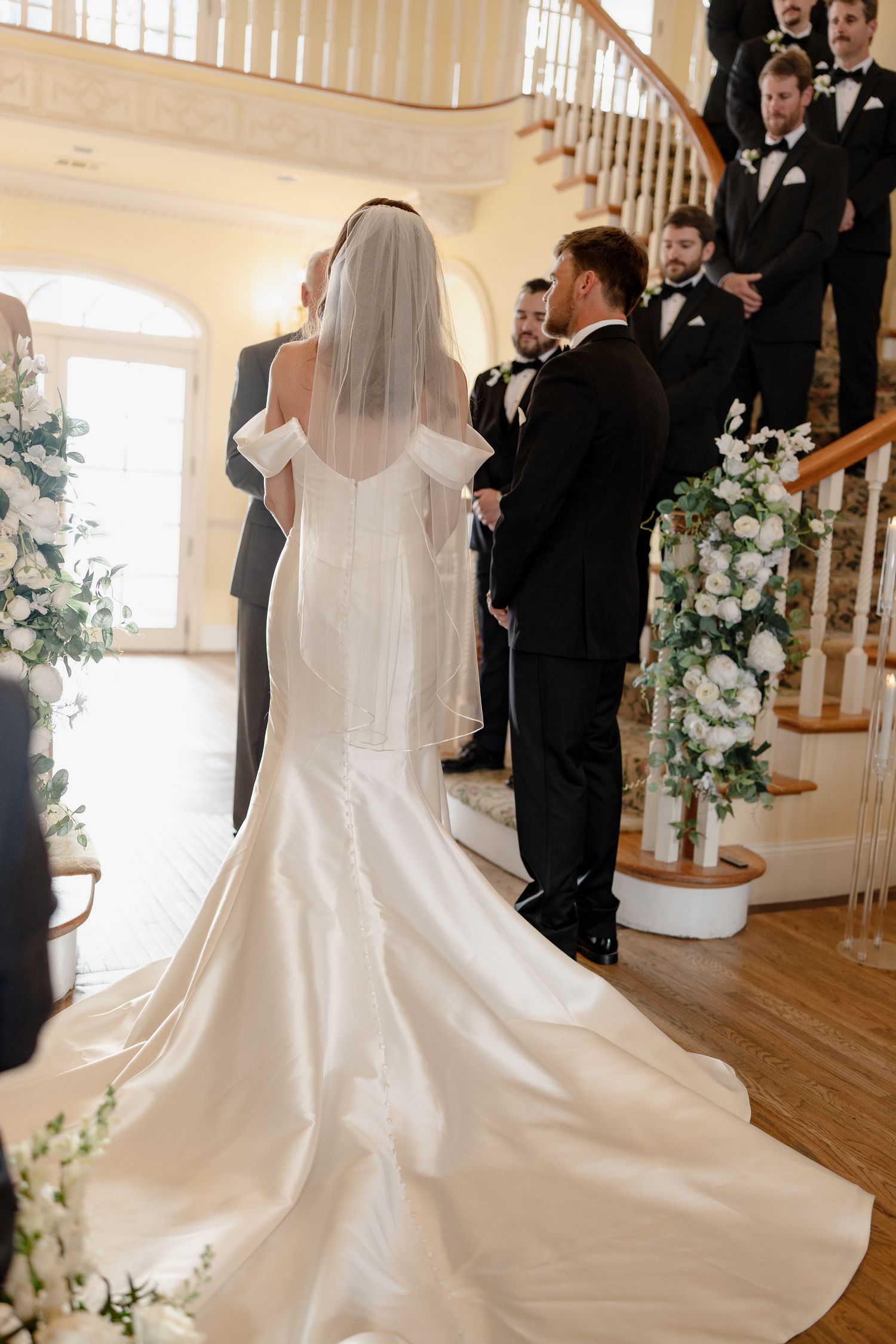 Bride in flowing white gown stands before wedding party during indoor ceremony.
