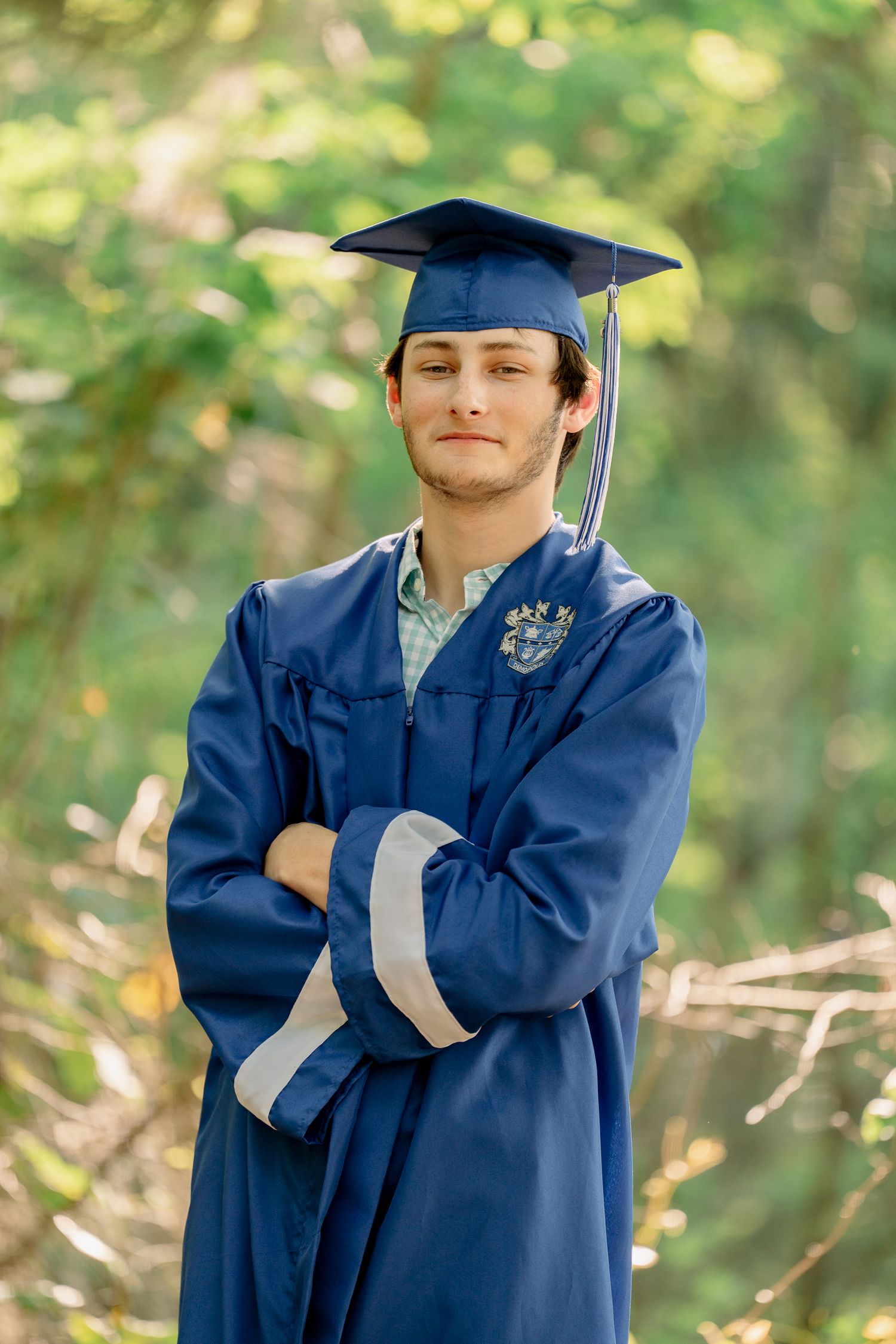 A graduate in a blue cap and gown posing outdoors with crossed arms and a content expression.