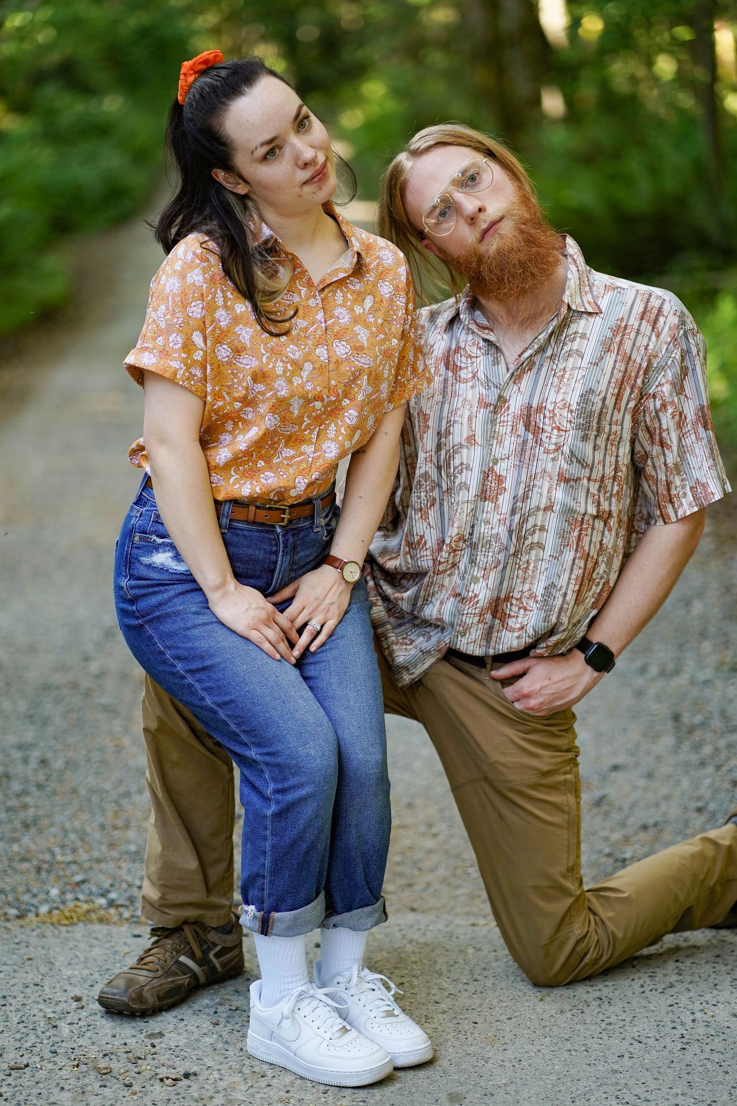 A couple in casual vintage-style clothing poses playfully on a forest path.