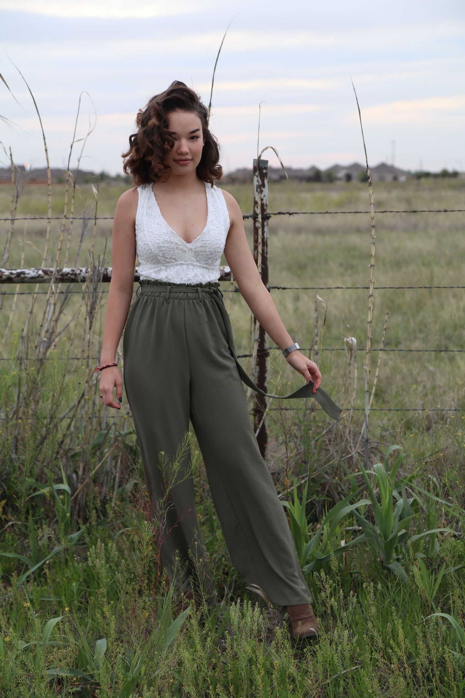 Woman in white lace top and olive pants poses by rustic fence in countryside setting.