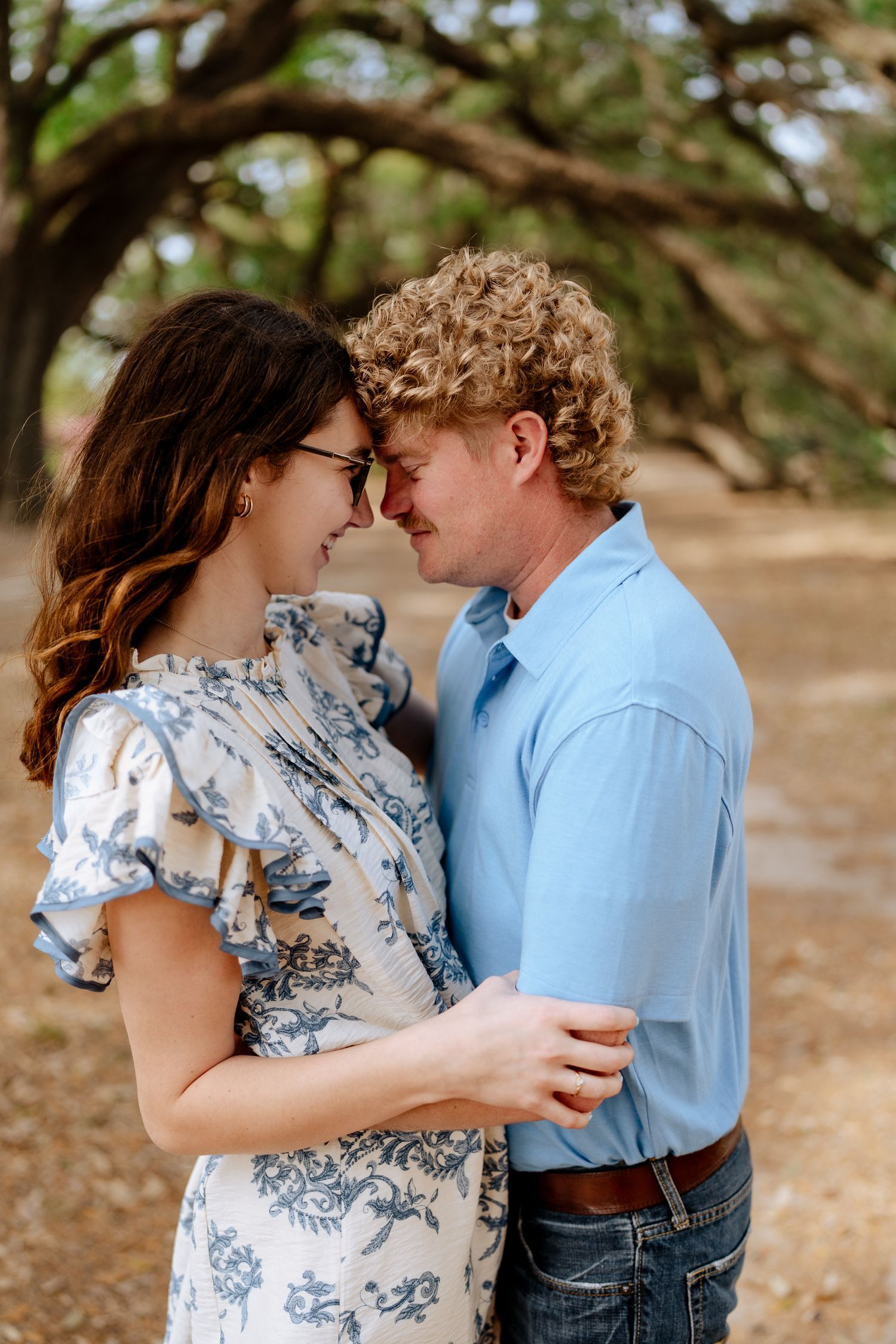 Couple in blue and floral outfits share romantic moments under trees.