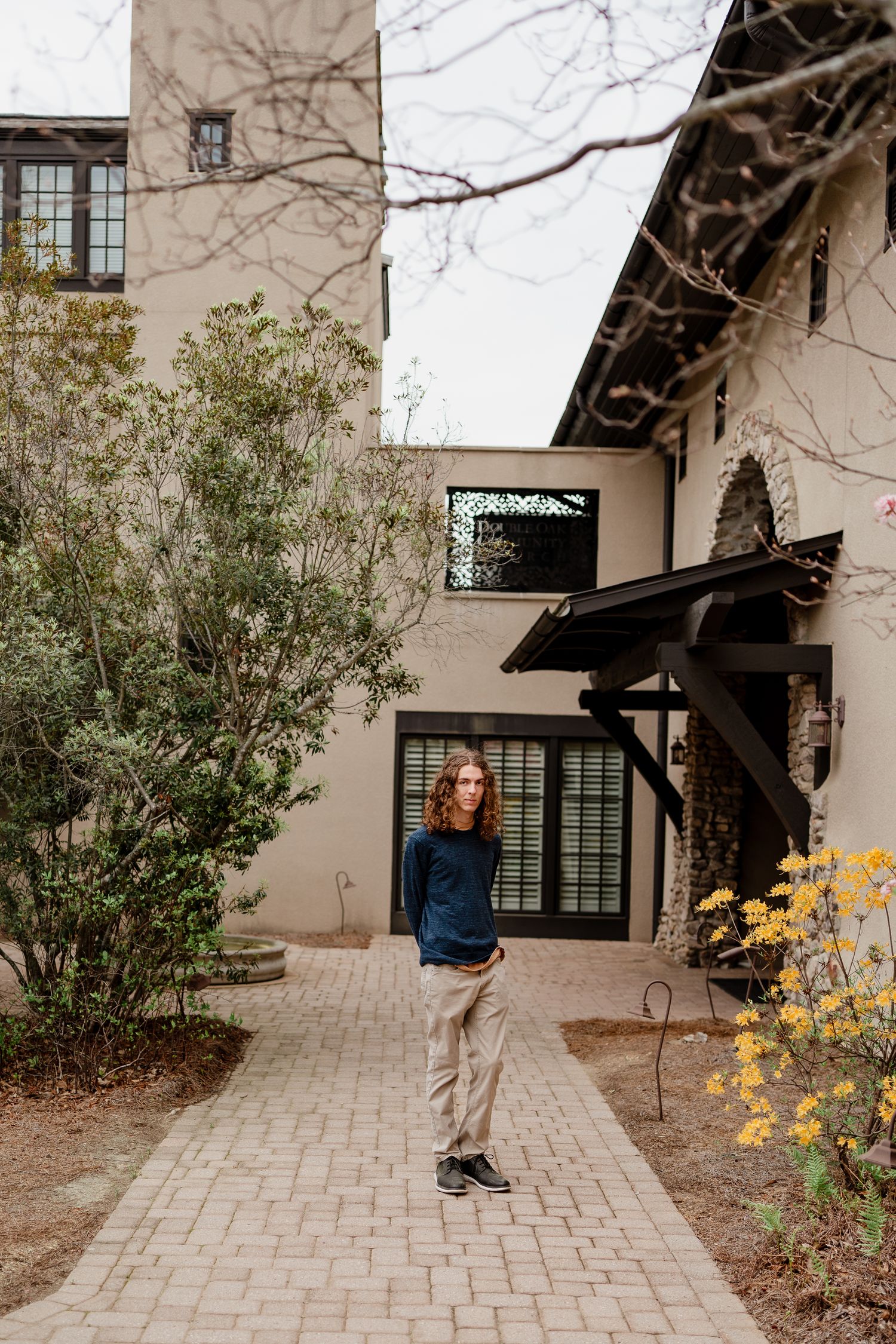 Person in blue sweater standing on brick pathway near beige building with landscaping.