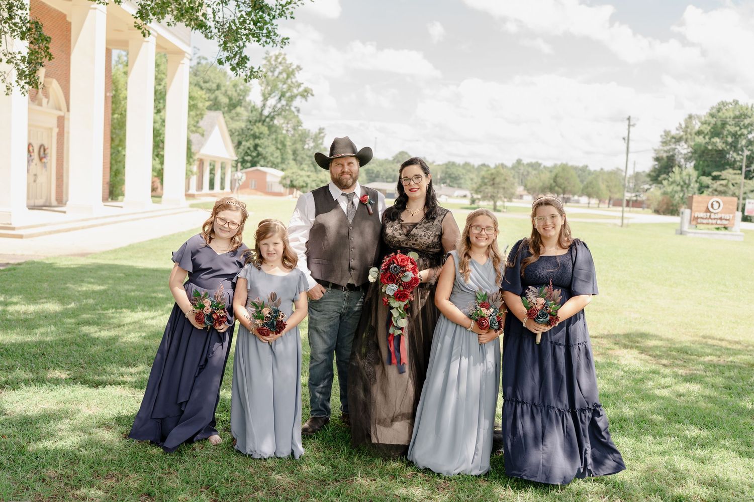 Wedding party in navy and gray dresses laughing together on a grassy lawn during an outdoor celebration.