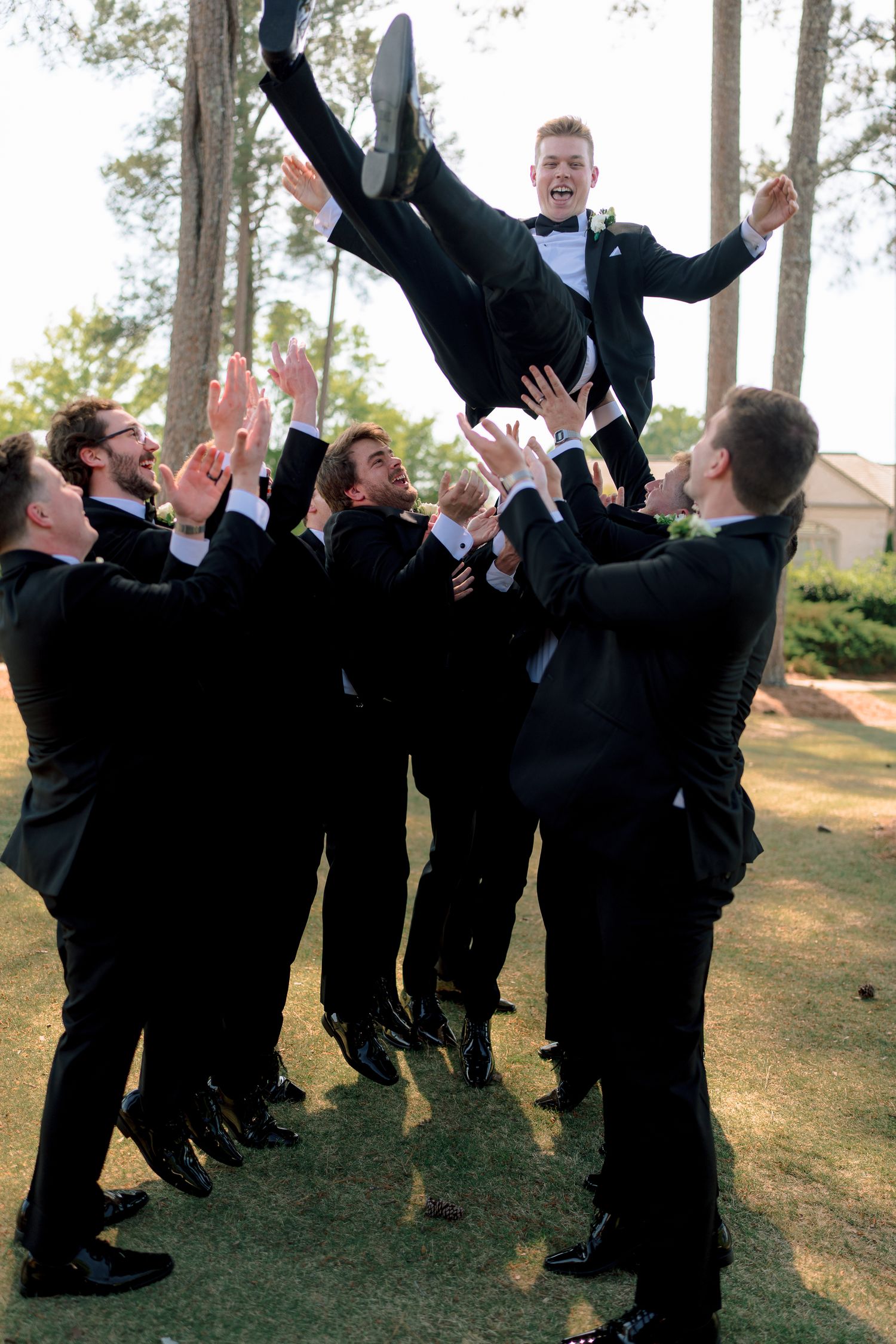 Groomsmen in black tuxedos tossing the groom into the air outdoors, celebrating on a sunny wedding day.