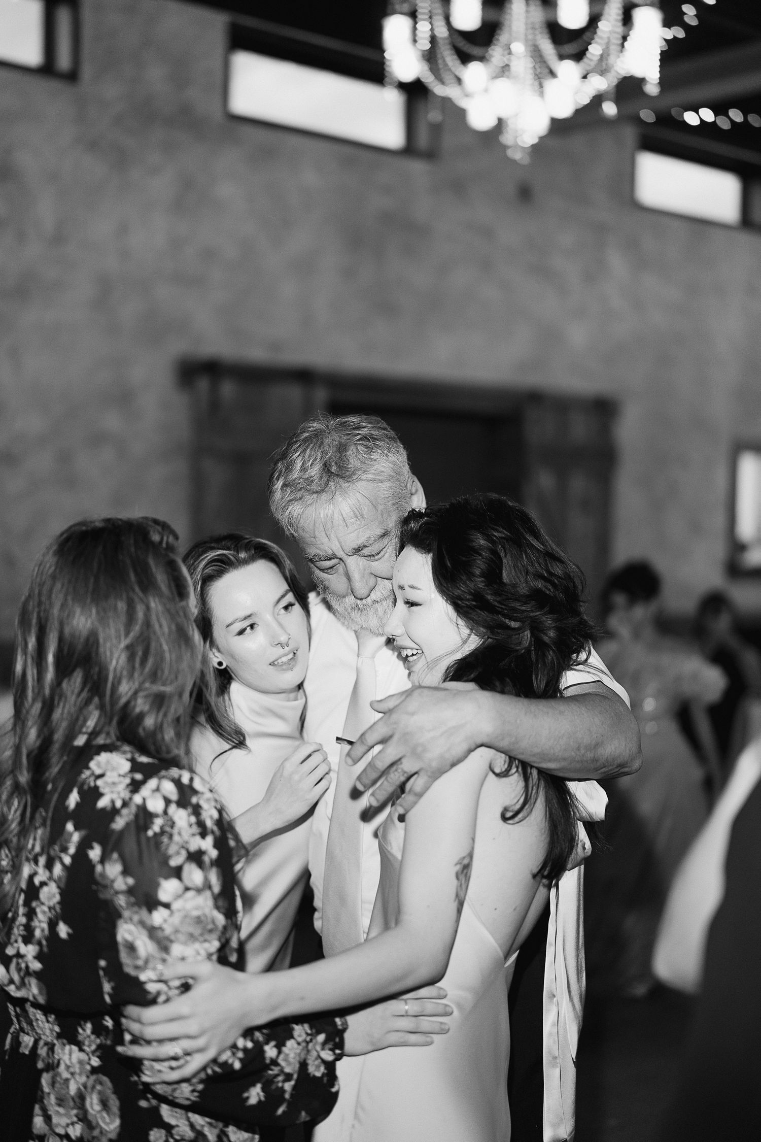 Black and white photo of a man and three women sharing a warm group hug at a wedding reception.