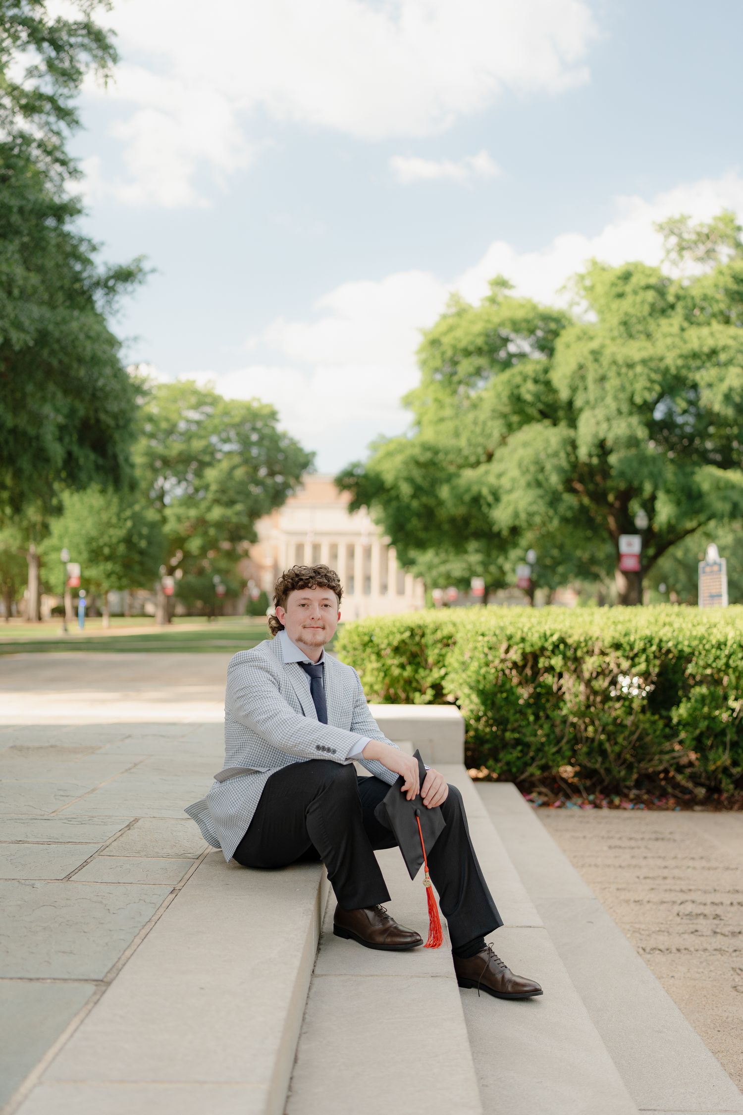 Young male graduate sits on steps holding diploma with red tassel on University of Alabama campus.