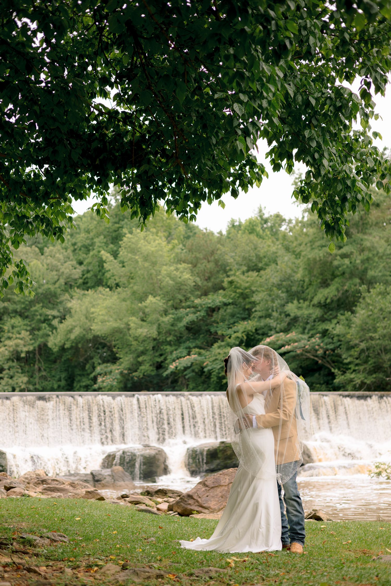 Bride and groom kissing under a veil beside a scenic waterfall surrounded by lush green trees.