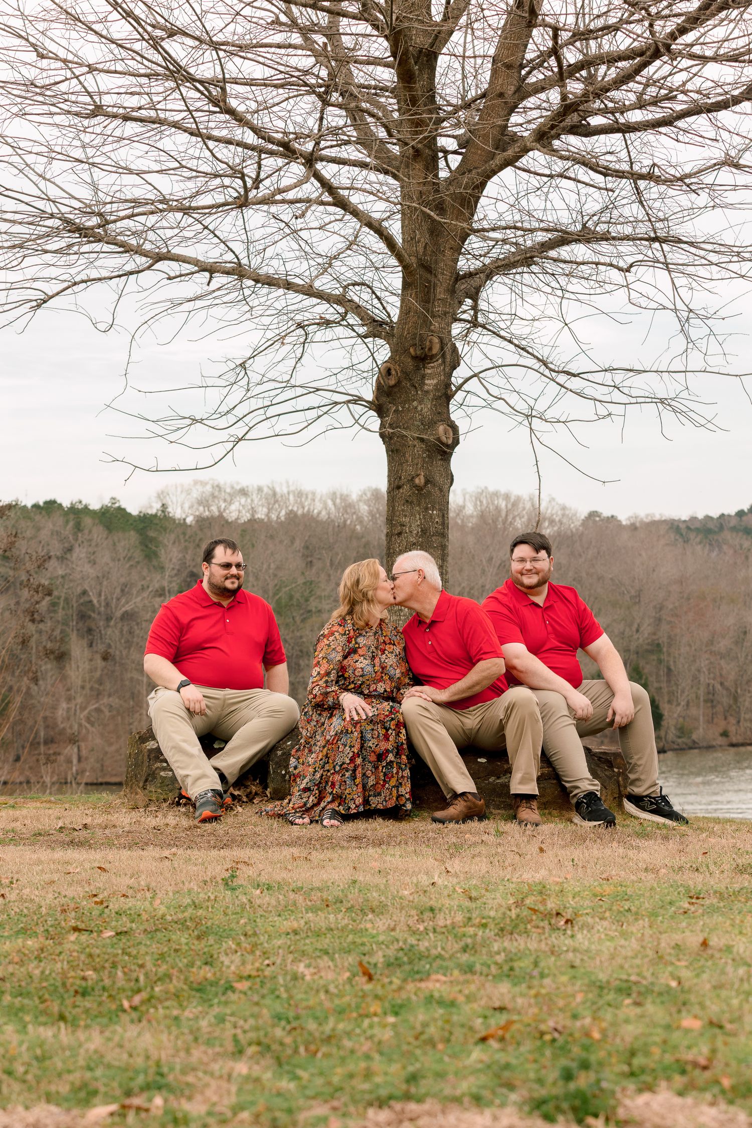 Family of four sitting under a bare tree near a lake, parents kissing while sons smile.