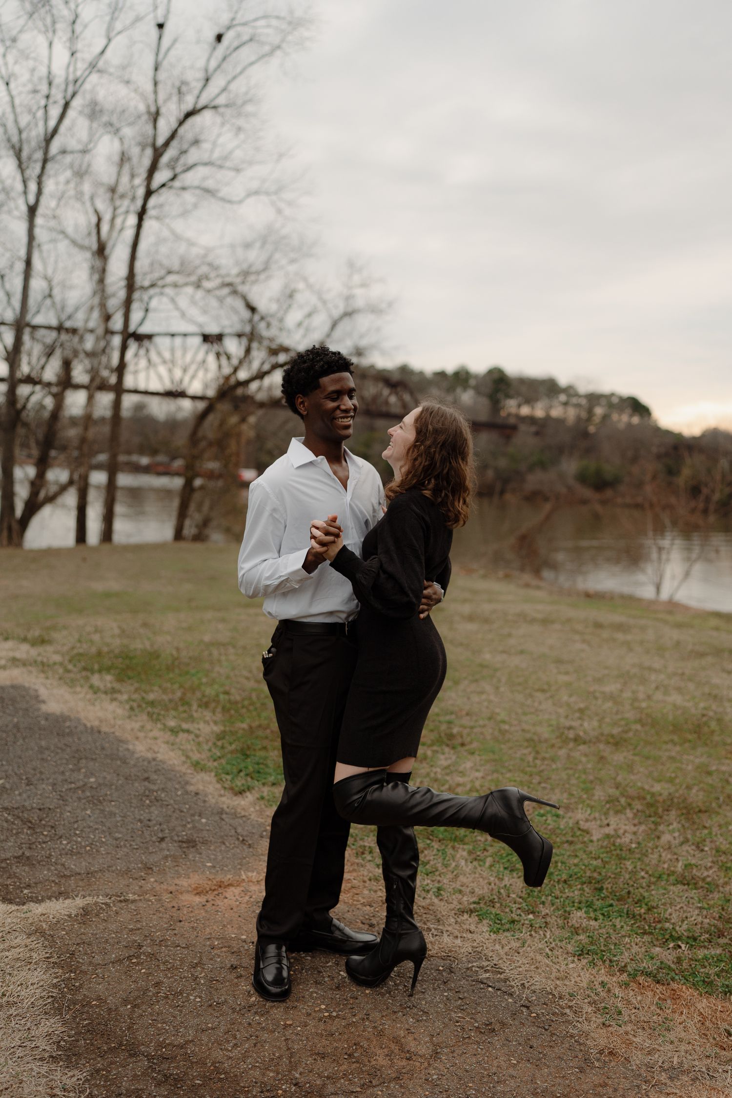 An interracial couple dances romantically outdoors near a river, the woman wearing knee-high boots and a black outfit.
