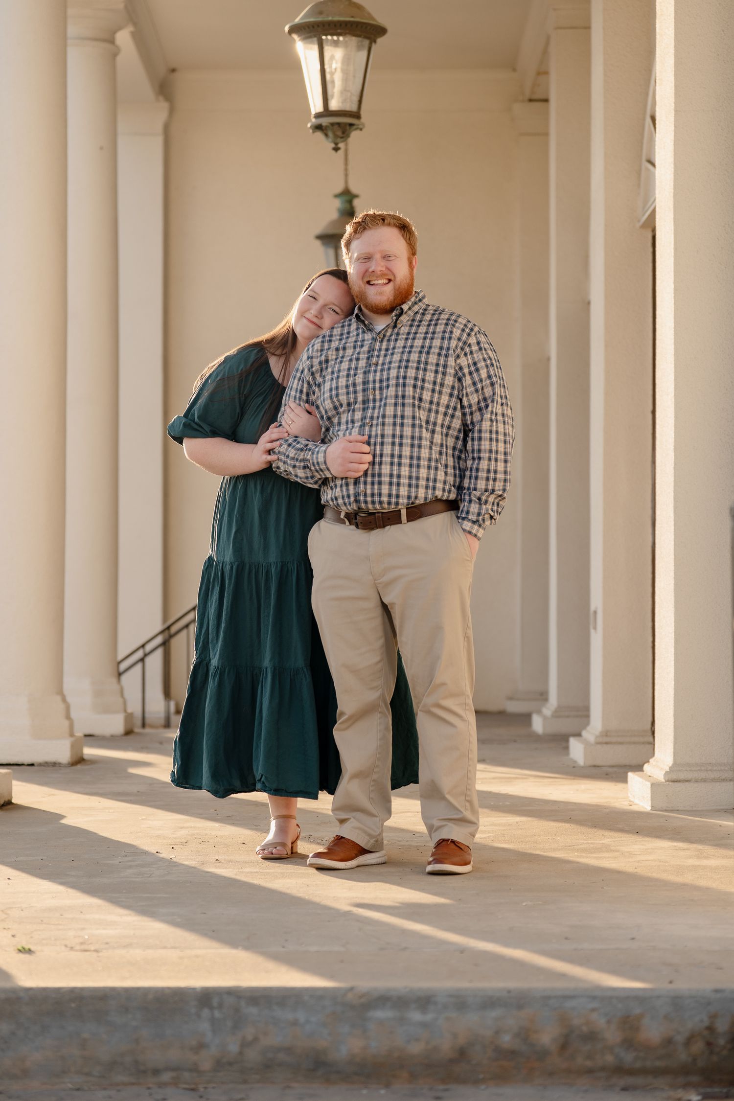 Happy couple embracing and laughing together under lantern between white columns in warm sunlight.
