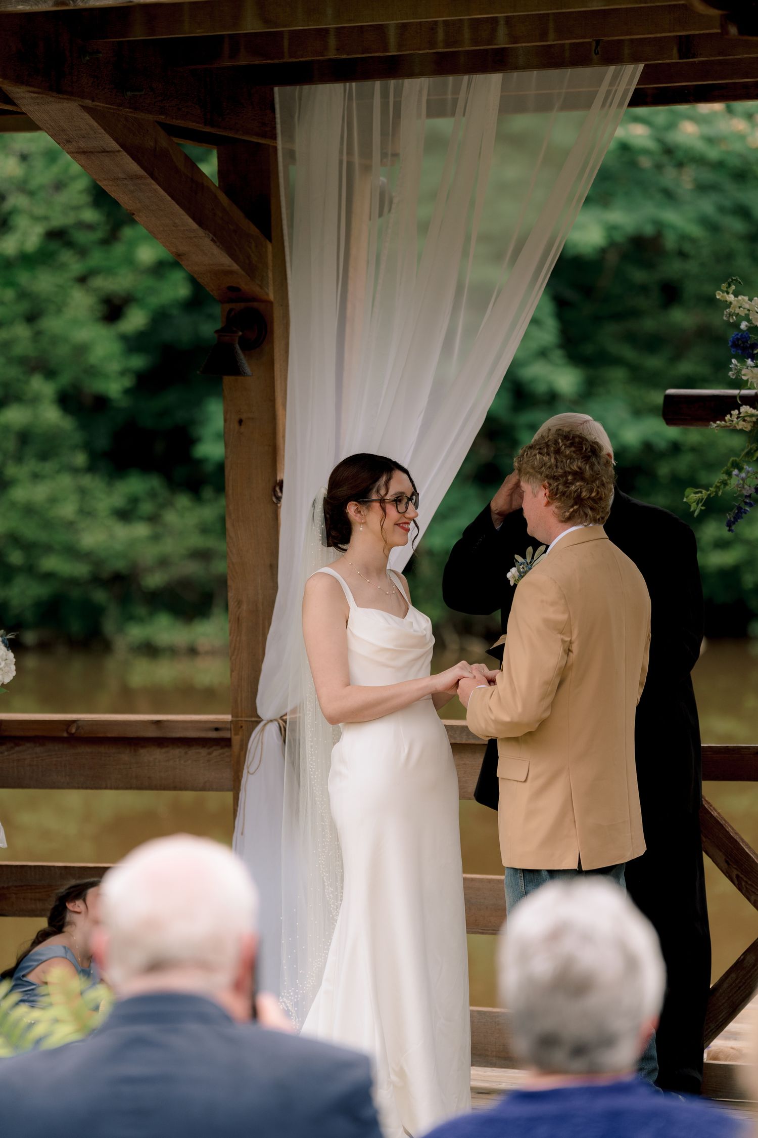 Bride in white gown and veil exchanges vows with groom in tan blazer at outdoor wedding ceremony by a river.