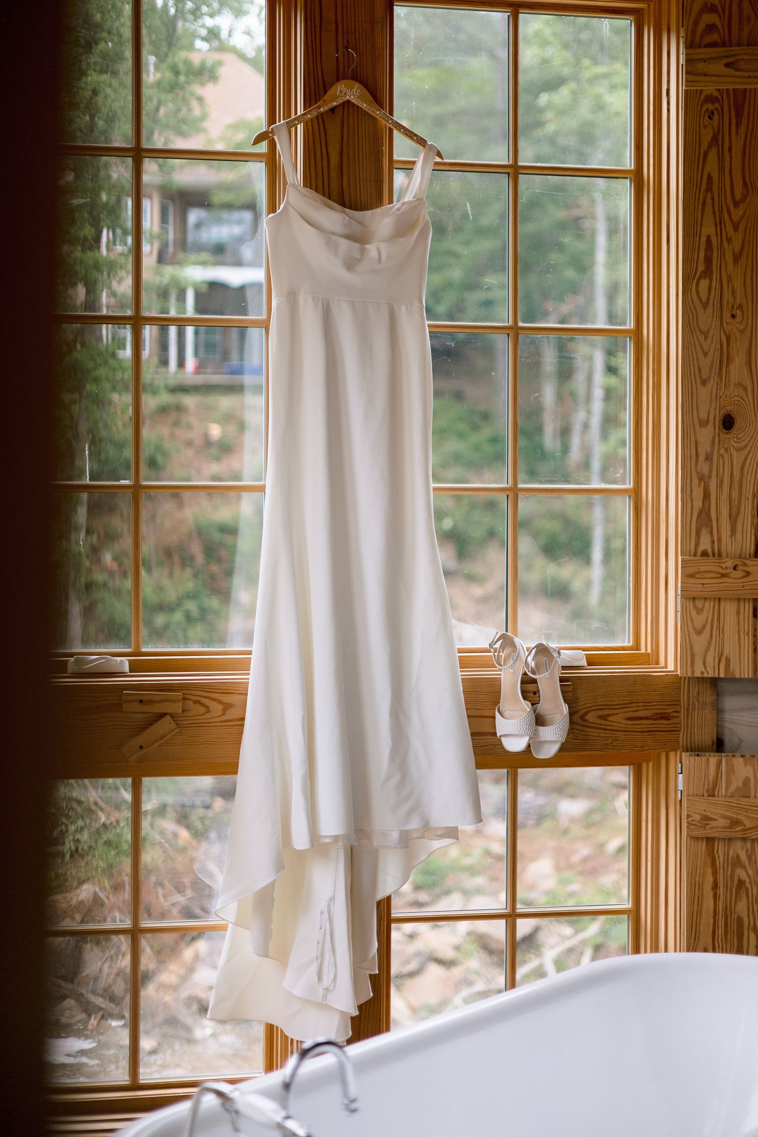 White wedding dress hanging on wooden window frame with white heels on sill above a freestanding bathtub.
