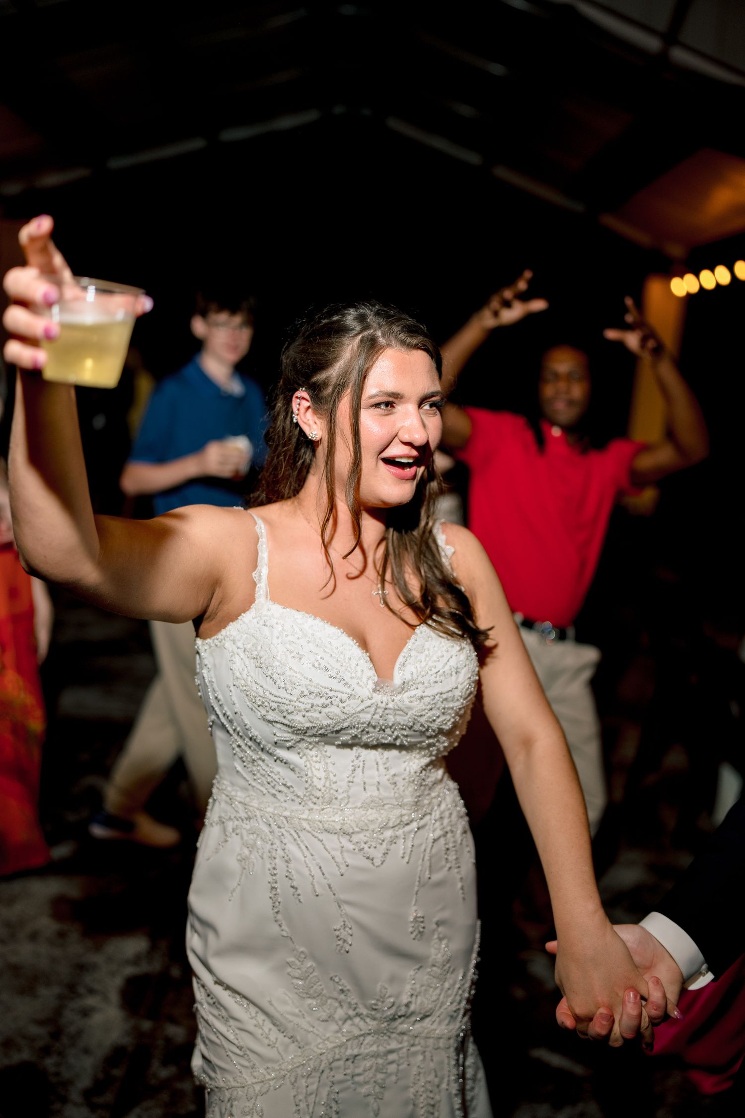 Joyful bride in lace gown dancing and raising a drink at lively wedding reception.