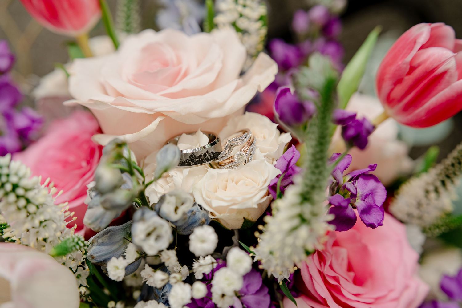 Close-up of a colorful wedding bouquet featuring pink roses, tulips, purple flowers, and two gold rings nestled among the blooms.