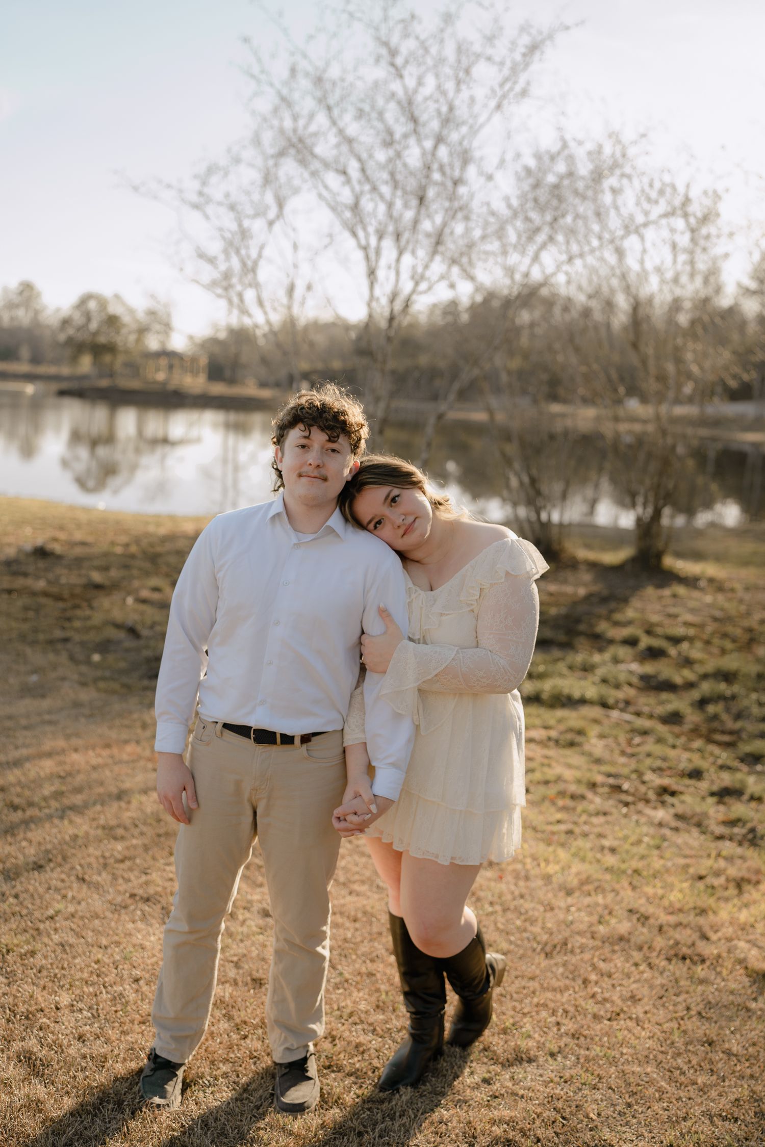 Couple standing close together by a lake at sunset, woman resting head on mans shoulder.