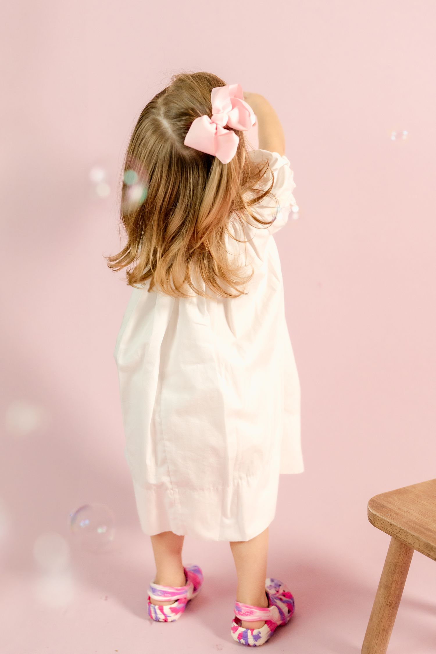 Little one in white dress and pink slippers plays with wooden stool in pink studio setting.