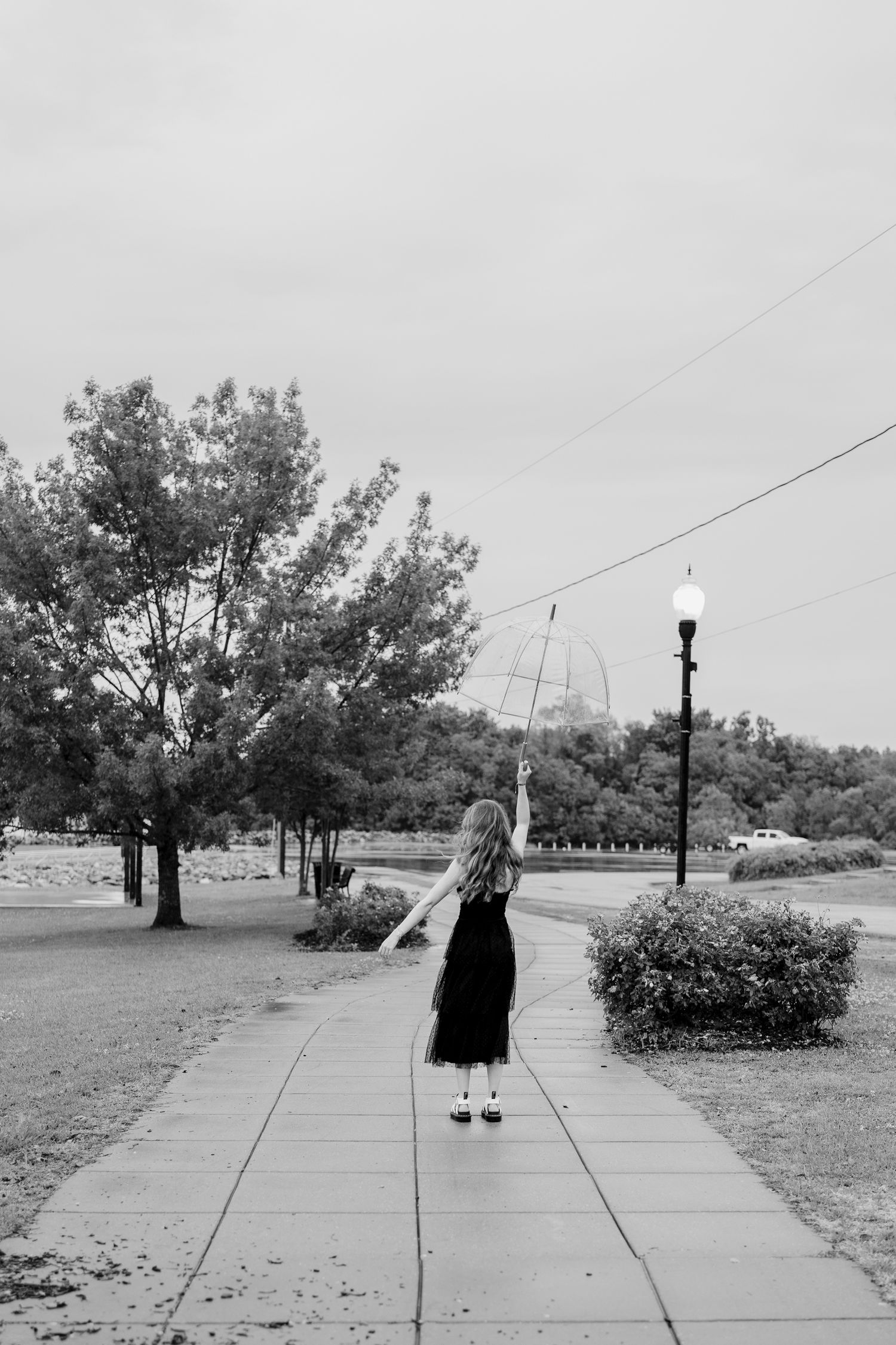 Black and white photo of someone in a dress holding an umbrella while walking down a tree-lined path.