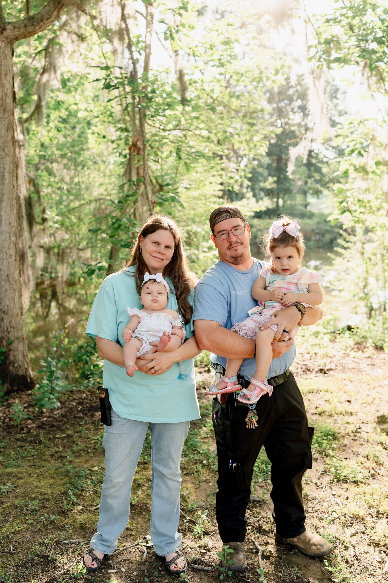 A family in matching light blue and black outfits poses together for outdoor photos in a sunlit wooded area.