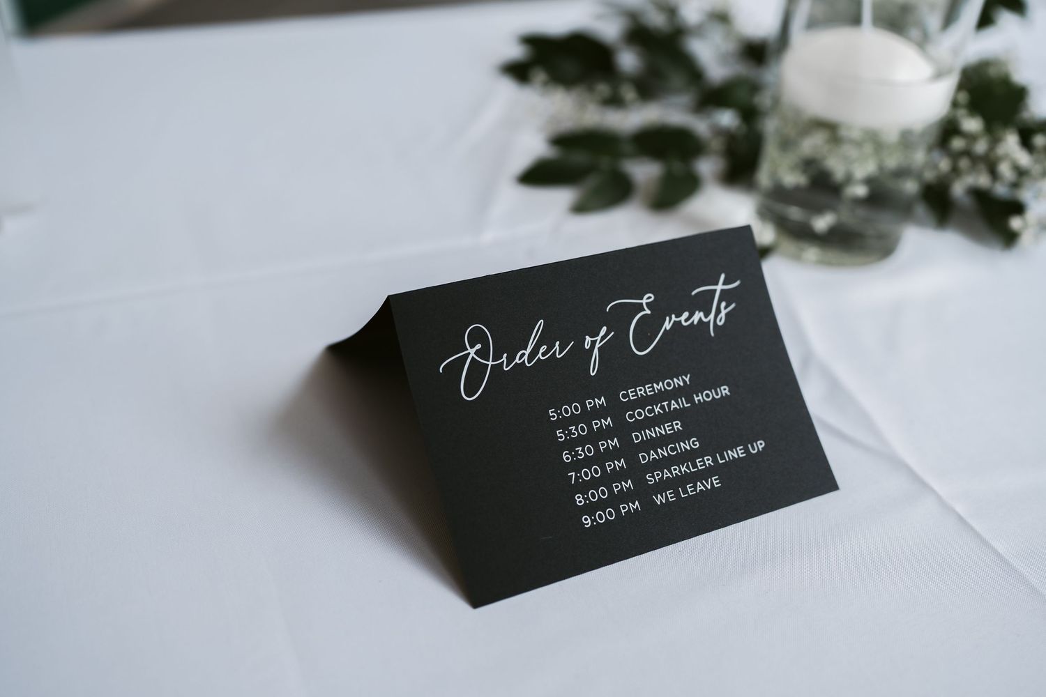 Black wedding event timeline card with white calligraphy on white linen tablecloth.