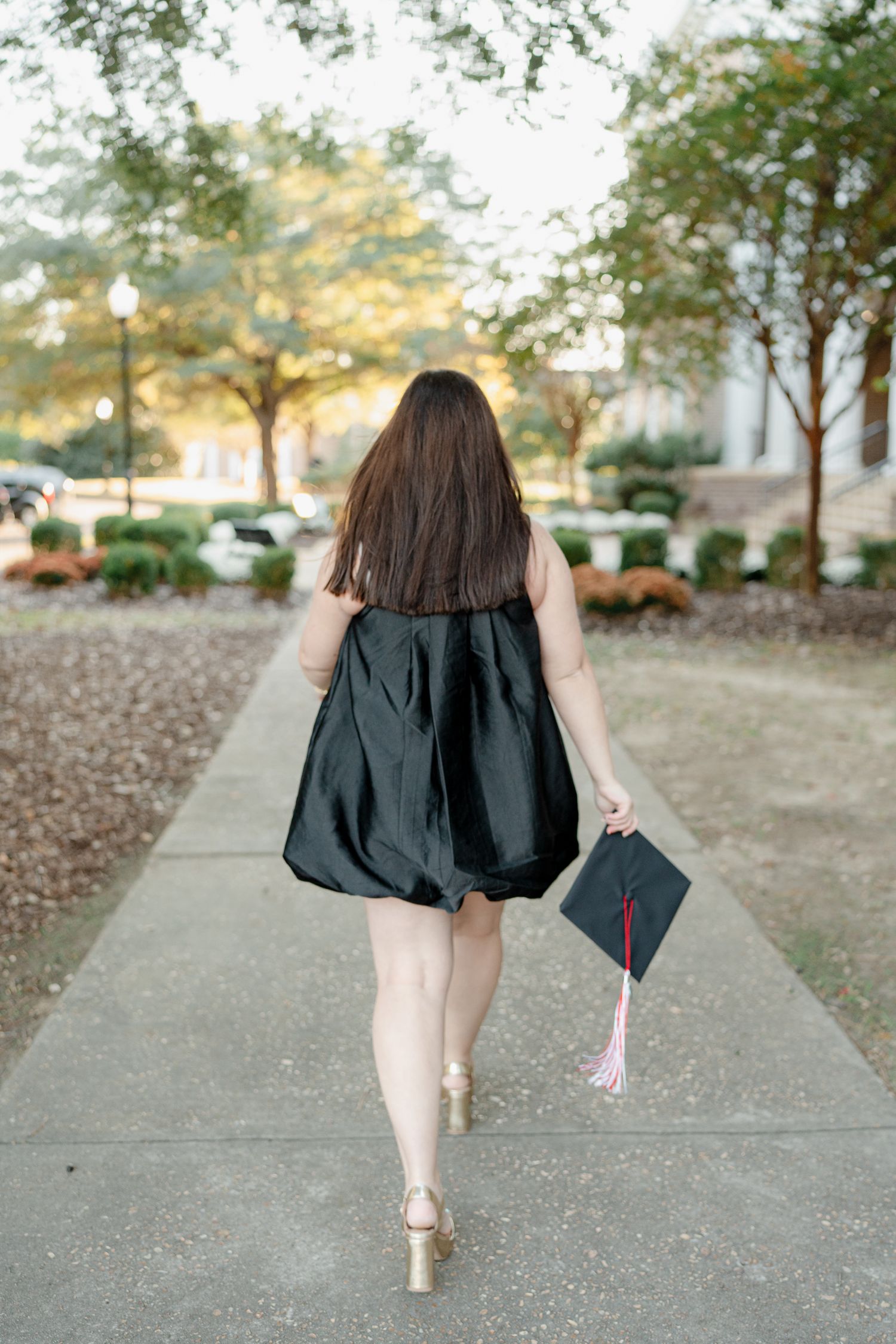A person in a black dress walks away on a concrete path while holding a graduation cap in their hand.