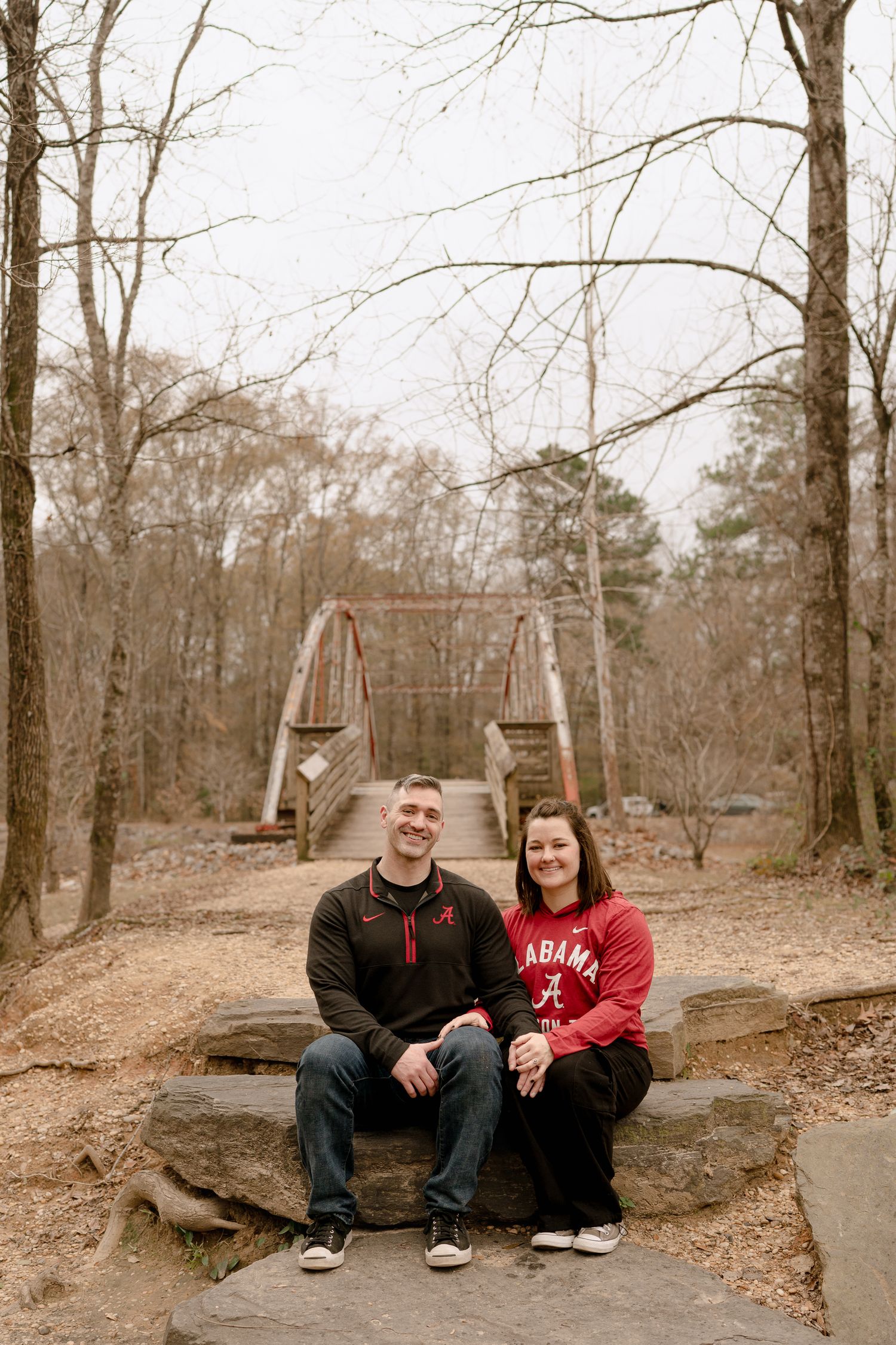 A couple sits together on a large rock in a wooded area during autumn.
