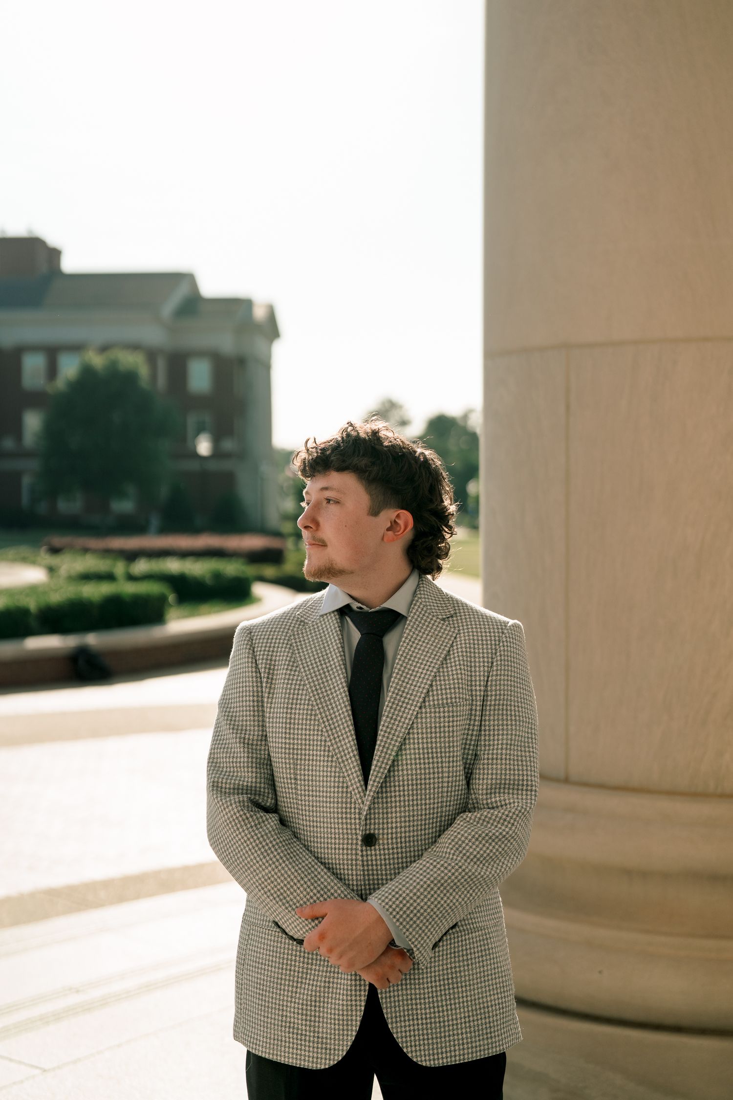 Young man in plaid blazer and tie stands beside a large column on a sunny college campus.