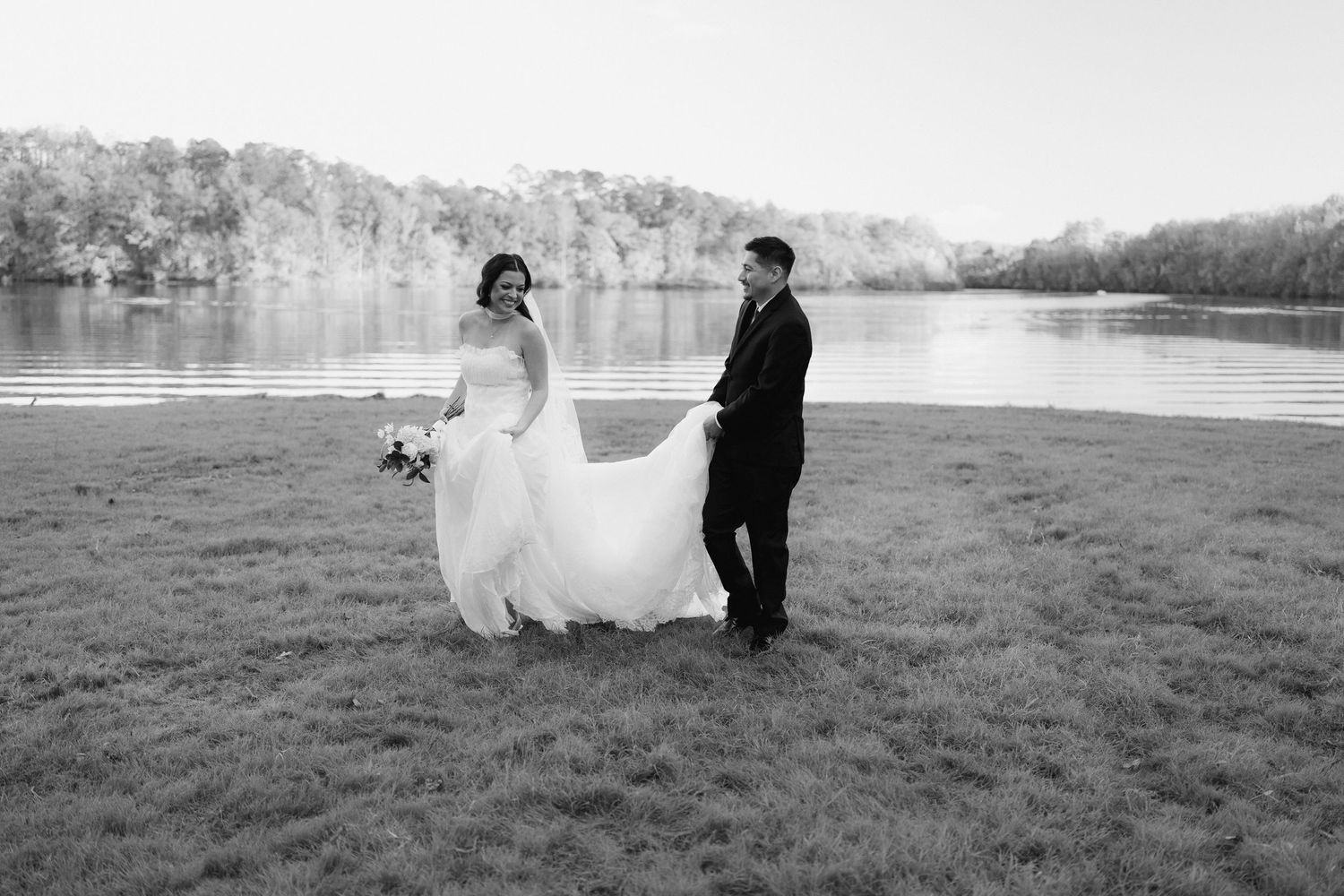 Black and white photo of a bride and groom standing by a lake, groom holding bride's wedding dress train.