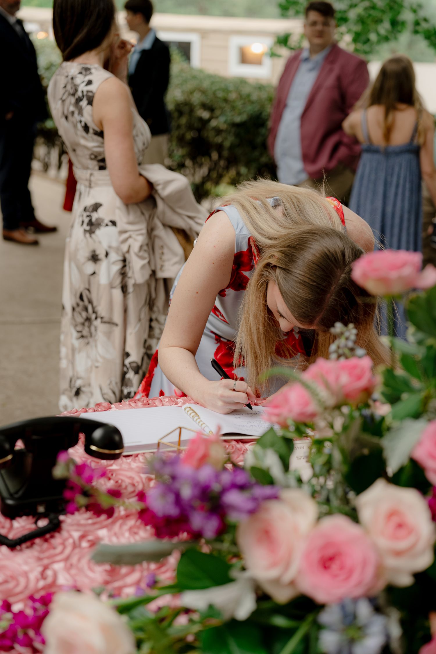 A wedding guest signs the guestbook at a floral-decorated table with pink roses in the foreground.