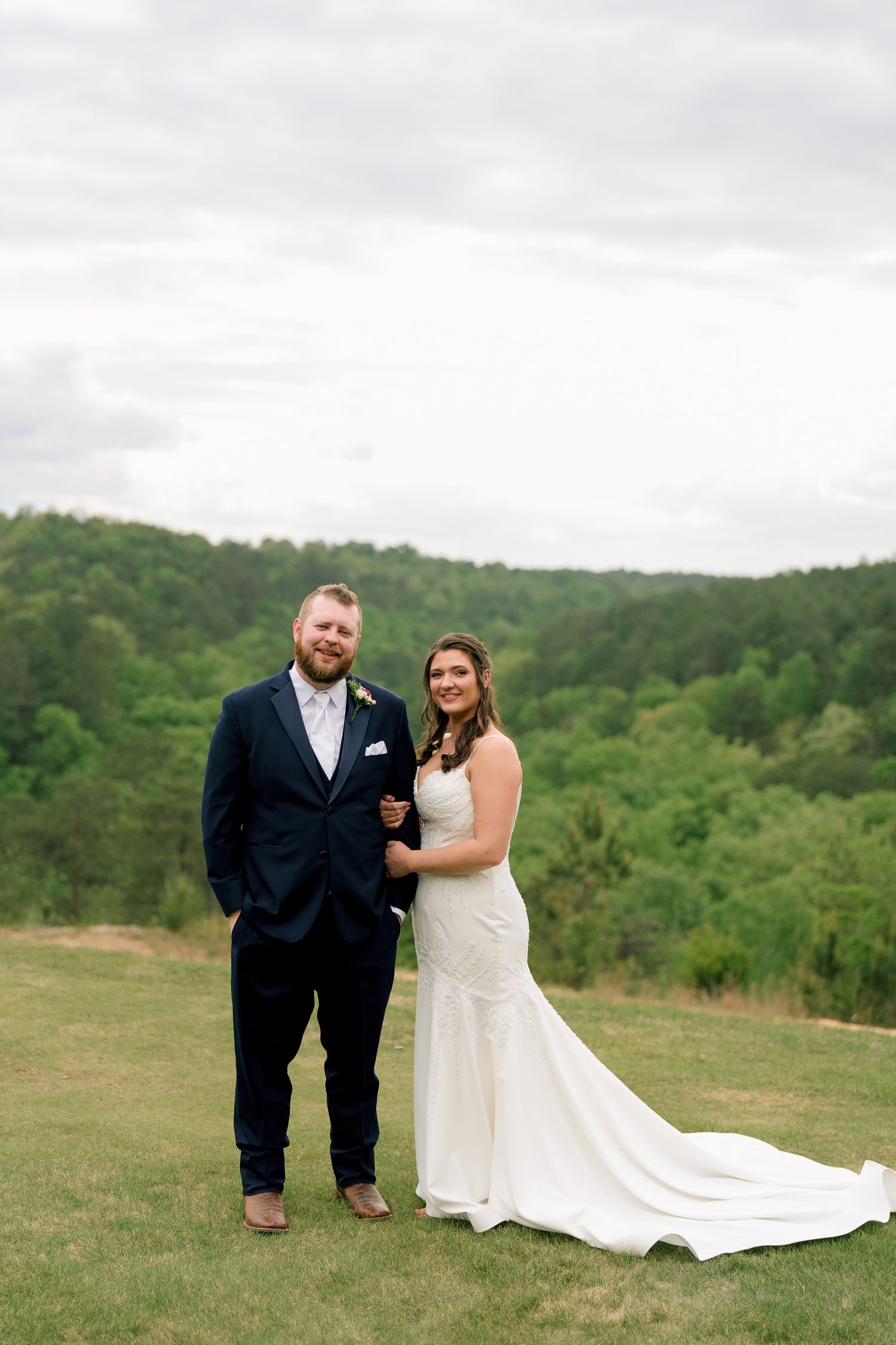 Bride and groom pose together on a green lawn with a forested hillside backdrop on their wedding day.