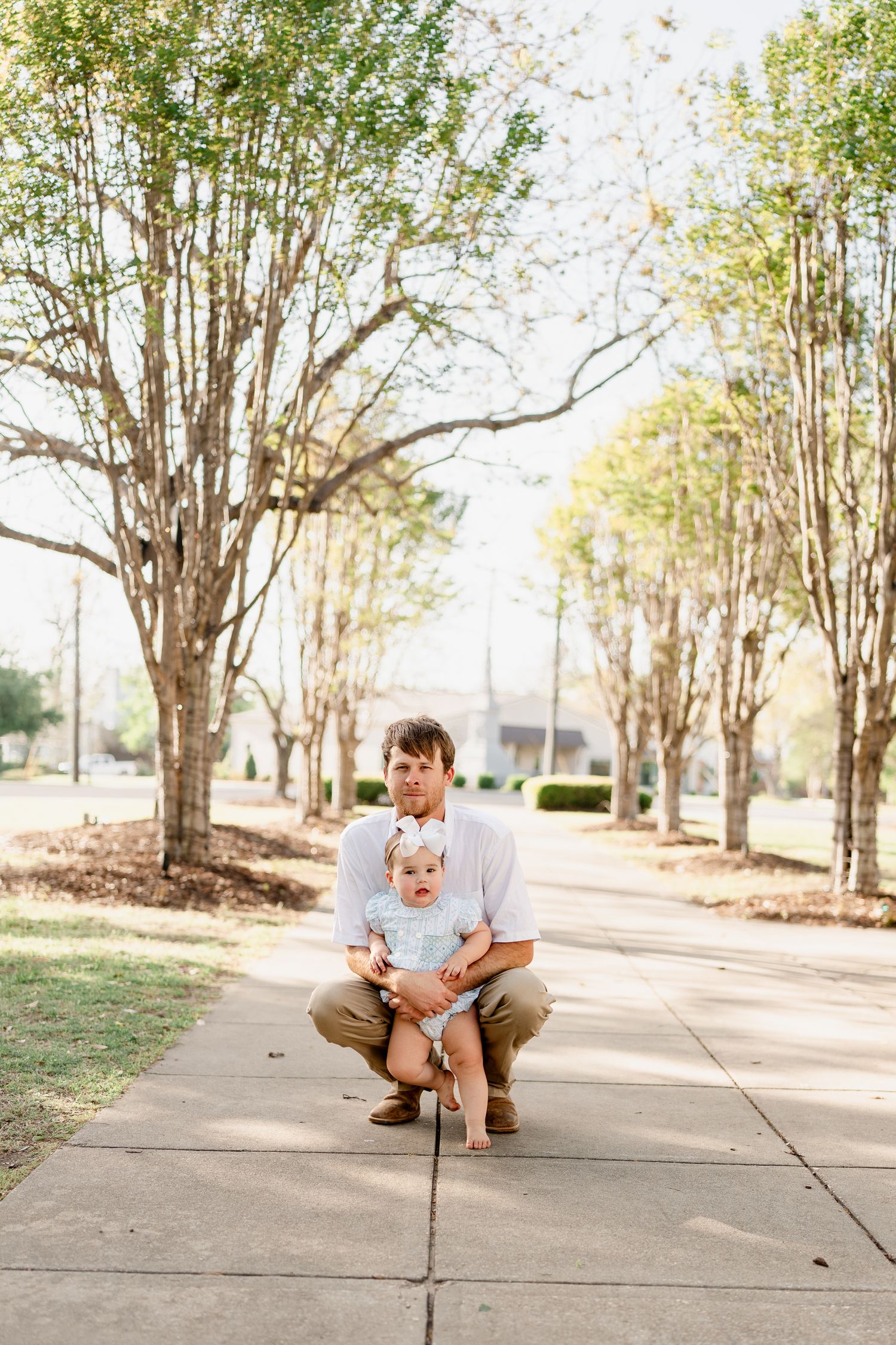 A parent crouches with a baby on a tree-lined walkway during a sunny outdoor portrait session.