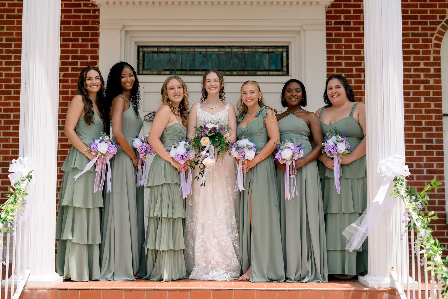 Bride and five bridesmaids in sage green dresses holding purple bouquets on brick steps.