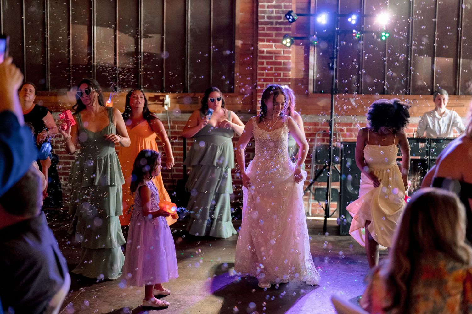 Bride in sparkly wedding gown dancing with guests and bridesmaids in sage dresses at reception.