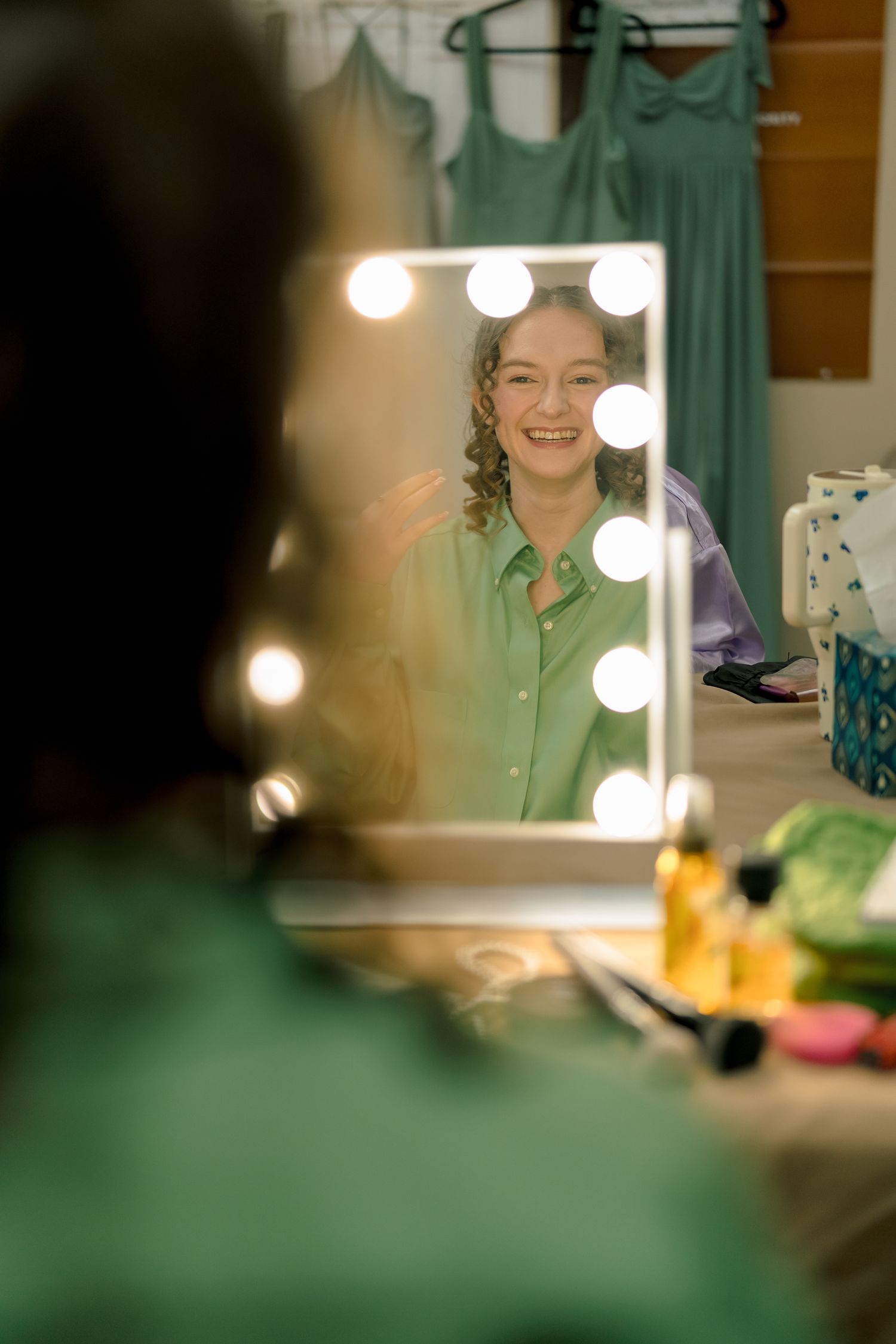Smiling bride applying makeup at a Hollywood mirror with lights in a green wedding preparation room.