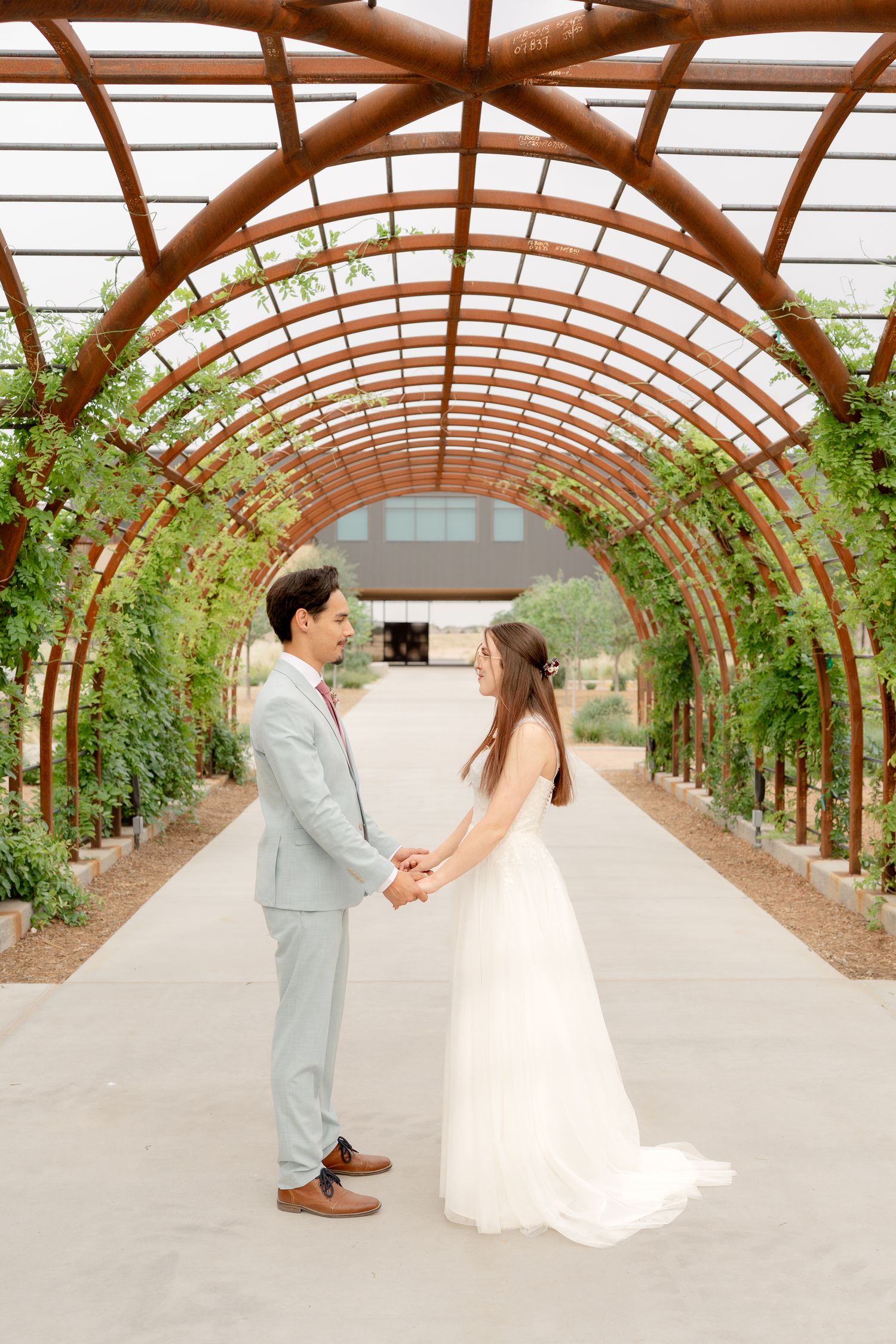 Couple shares a romantic moment in a sunlit greenhouse walkway with arched wooden beams and climbing vines.