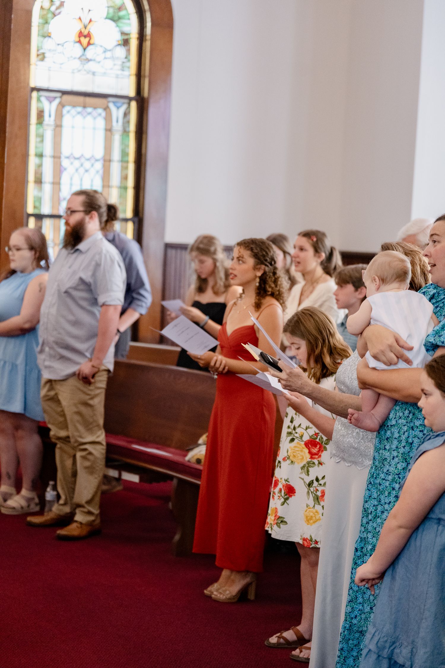 Wedding guests stand in church pews singing from hymnals during a ceremony with stained glass windows visible.