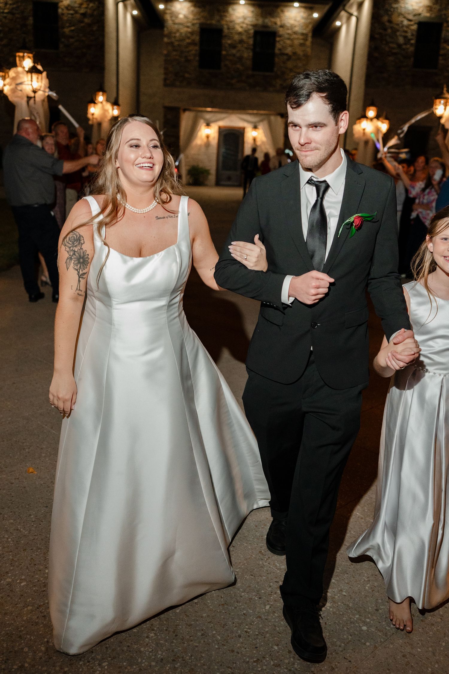 A bride in a white satin dress and groom in black suit walk together during an evening reception with warm lighting.