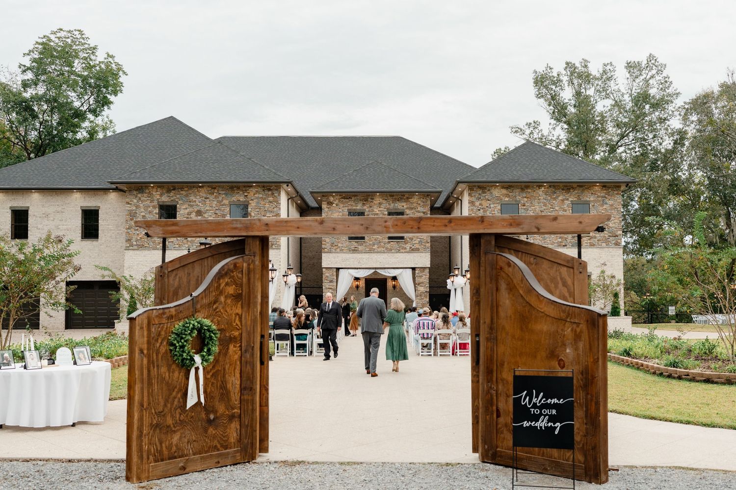 Rustic wooden gates open to an elegant stone venue with outdoor event setup and greenery in the background.