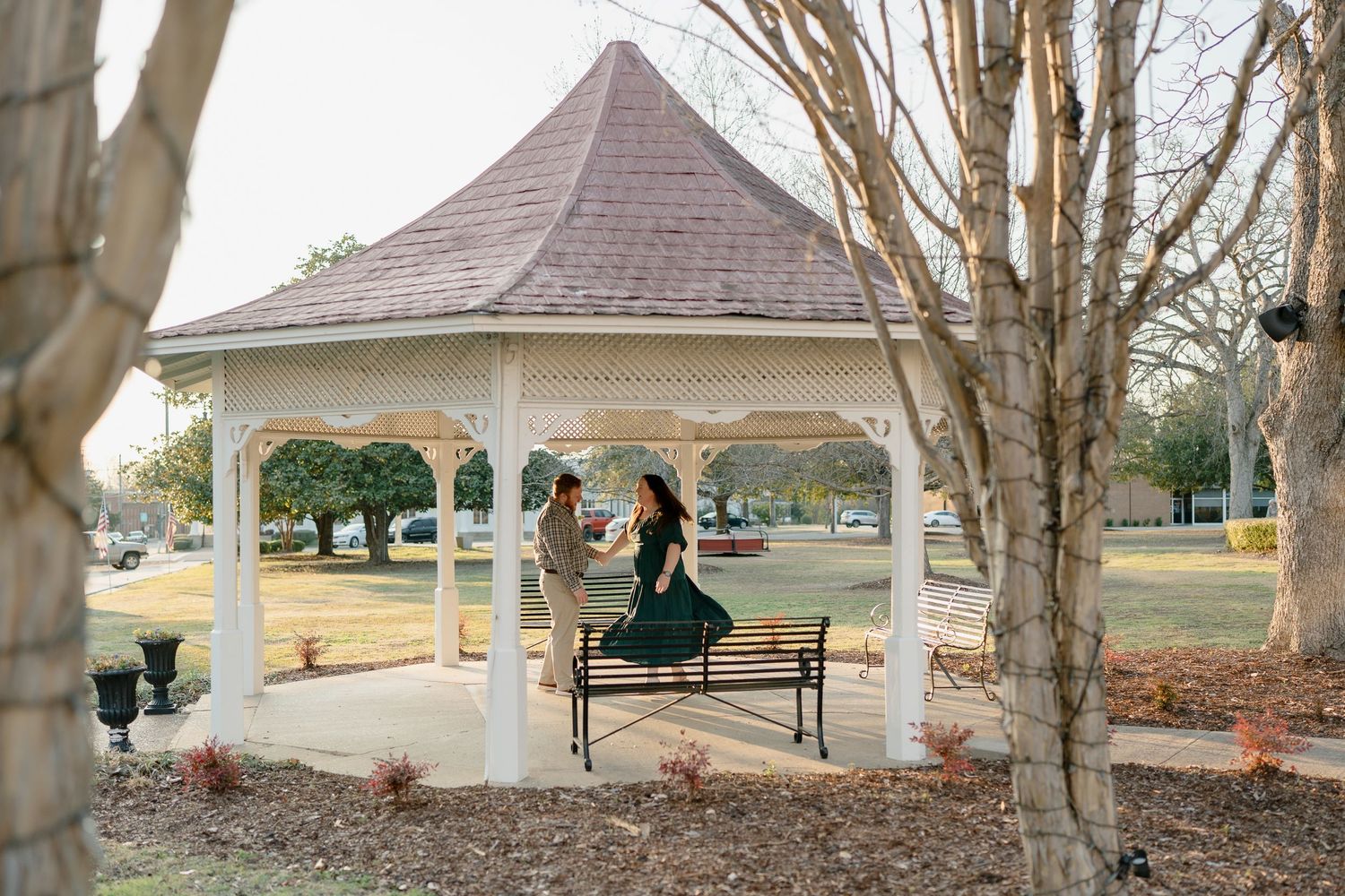 Couple sharing a romantic moment inside a white gazebo surrounded by trees in a park setting.